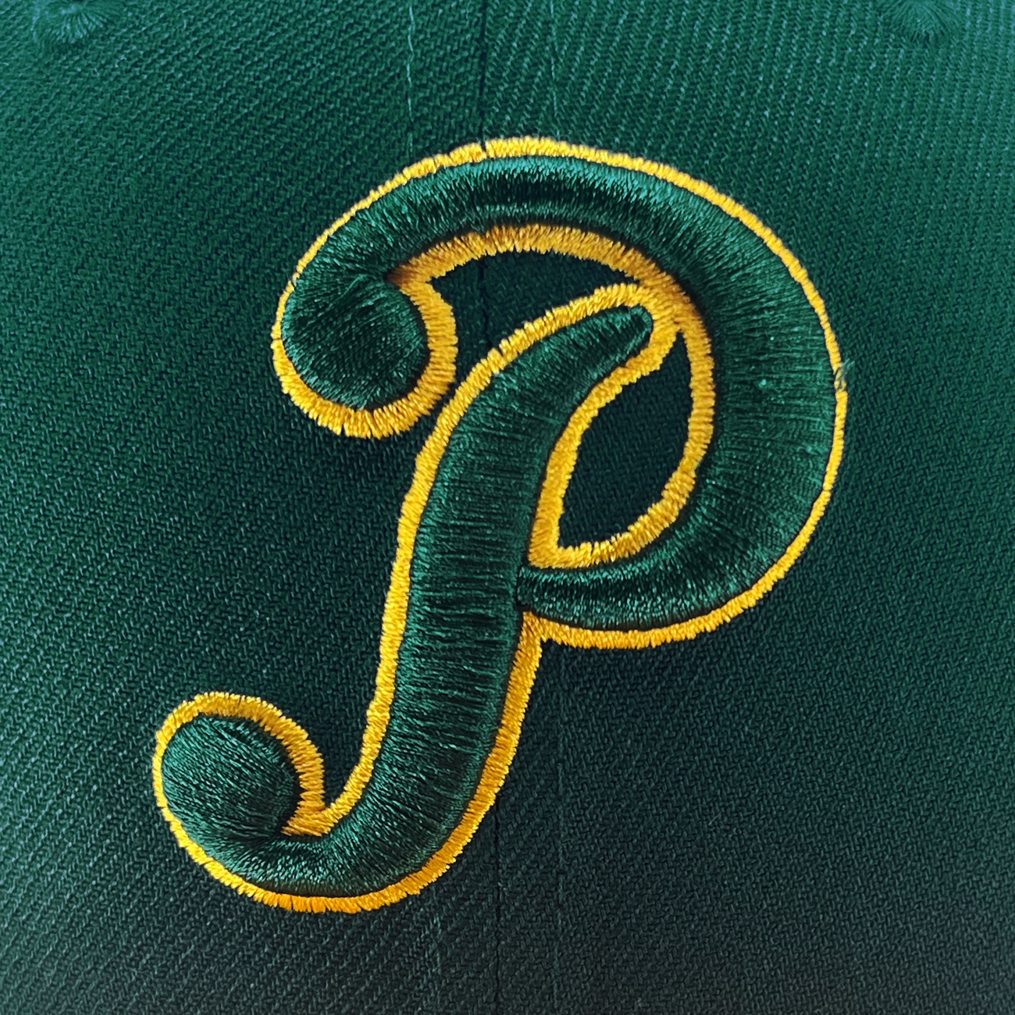 A close-up image of the dark green fabric on the New Era Pericos Home 59FIFTY Cap by Retail Vendors, featuring an intricately embroidered yellow and green letter "P" in a cursive style. The bold design of the "P" and the detailed stitching clearly showcase the texture and craftsmanship of the embroidery.