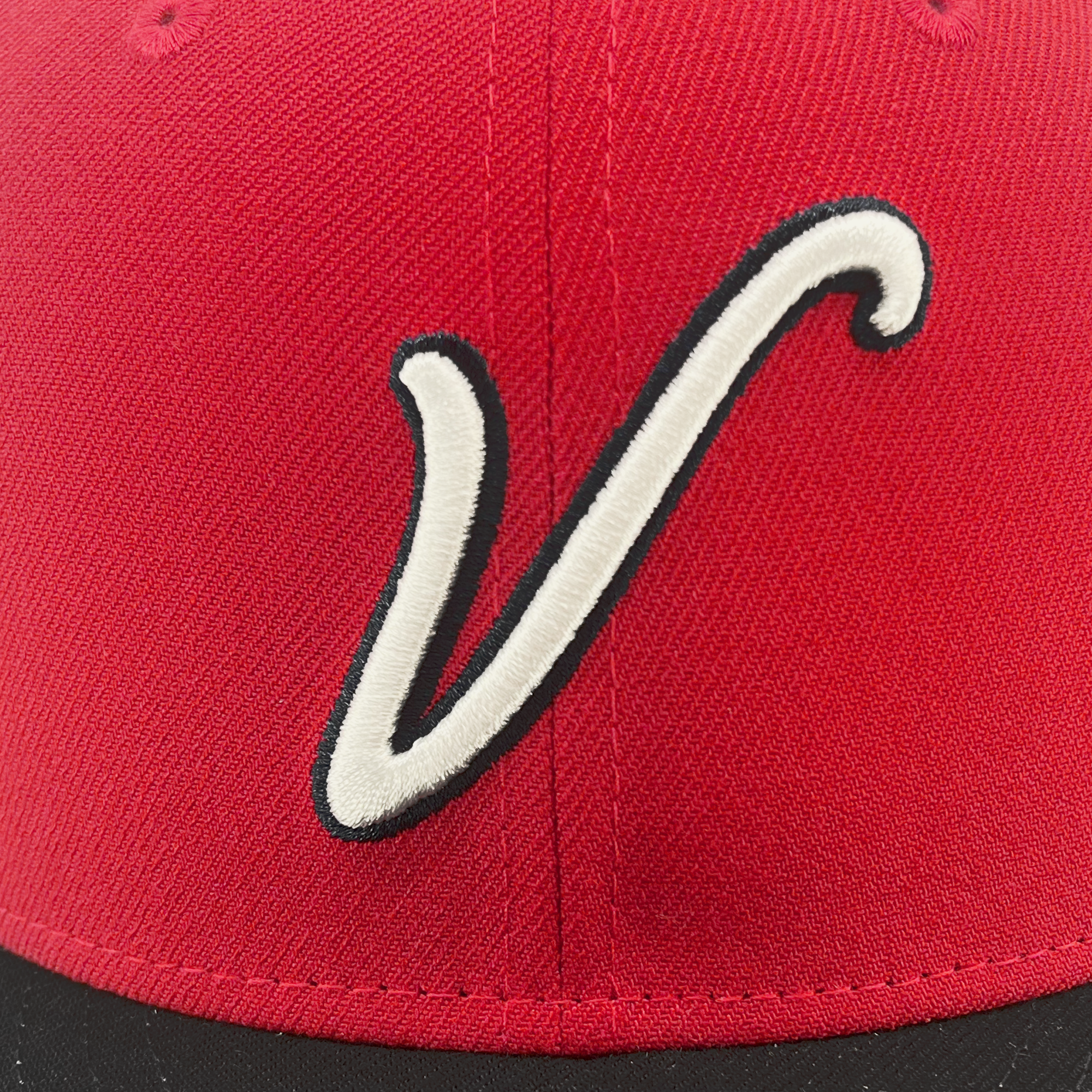 A close-up of the New Era El Aguila Alt 59FIFTY cap by Retail Vendors, showcasing a stylized white letter "V" outlined in black on the front. The red fabric's stitching and texture are clearly visible, with the bottom edge of the cap in black.