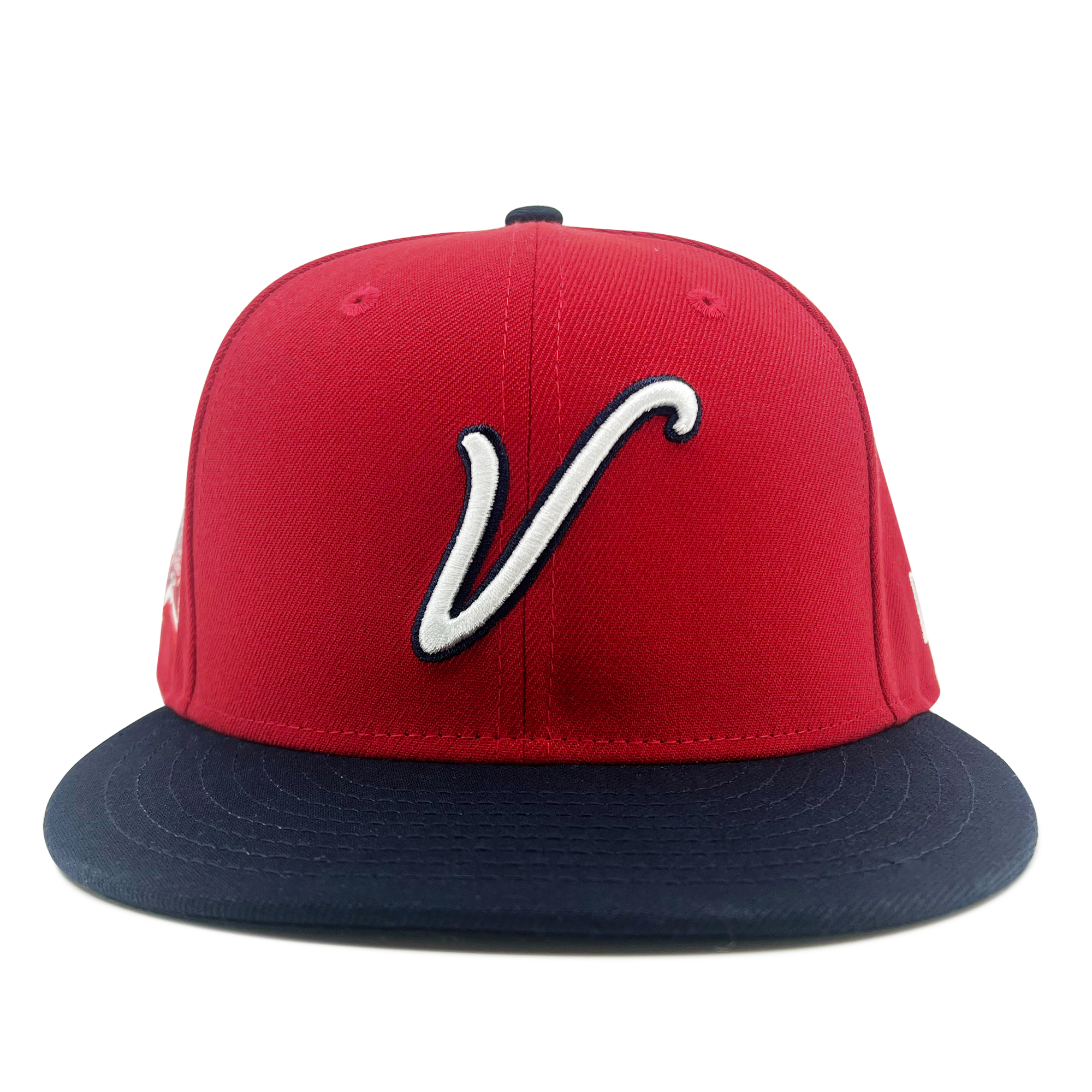 The New Era El Aguila Alt 59FIFTY Cap by Retail Vendors is a red baseball cap with a navy blue flat brim and stitched eyelets for ventilation. The front features a white stylized "V" logo, set against a plain white background that accentuates the cap's vibrant colors.