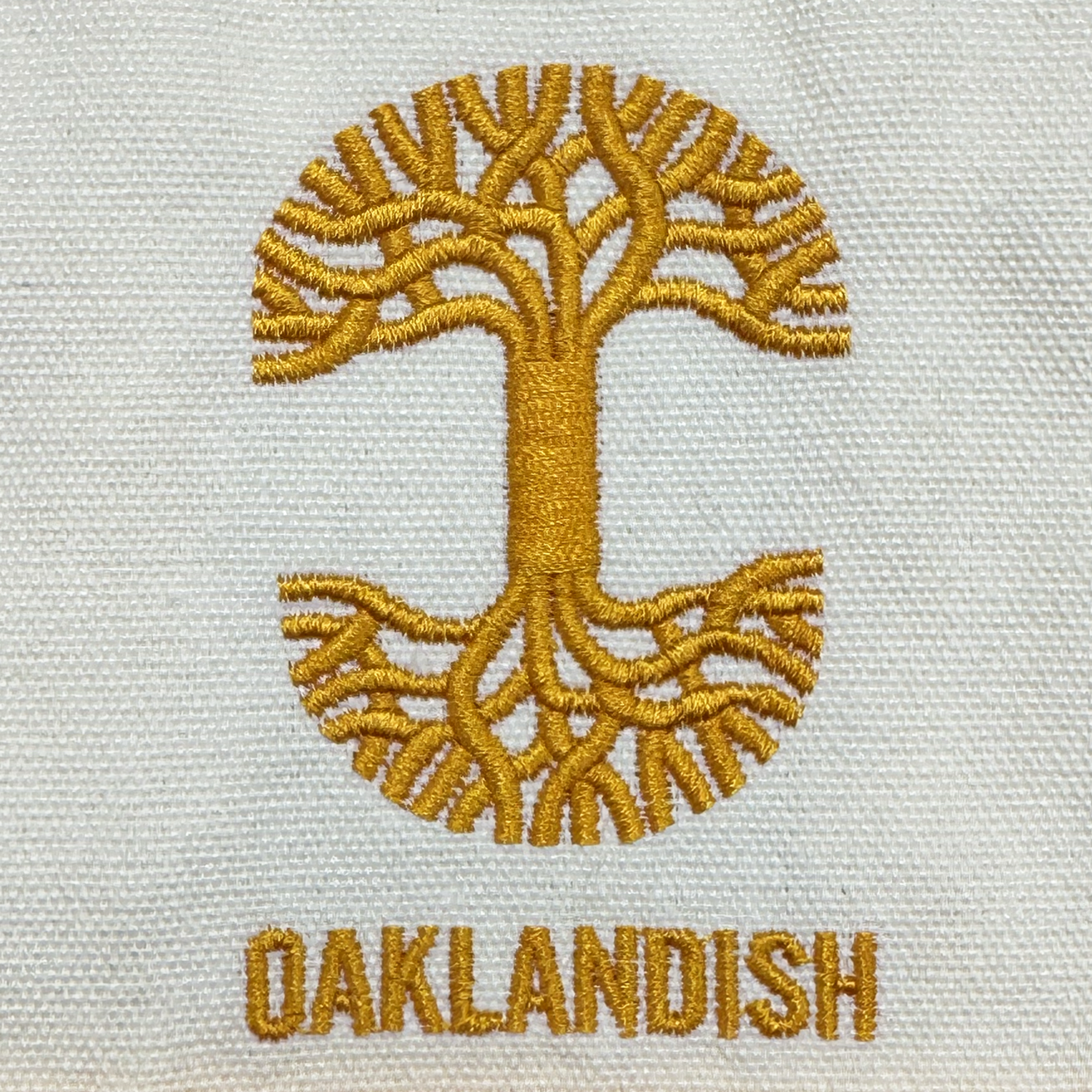 The Oaklandish Mini Grocery Tote features a gold embroidered tree with branching roots and canopy forming a circle on cream fabric, with "OAKLANDISH" stitched in gold capital letters beneath.