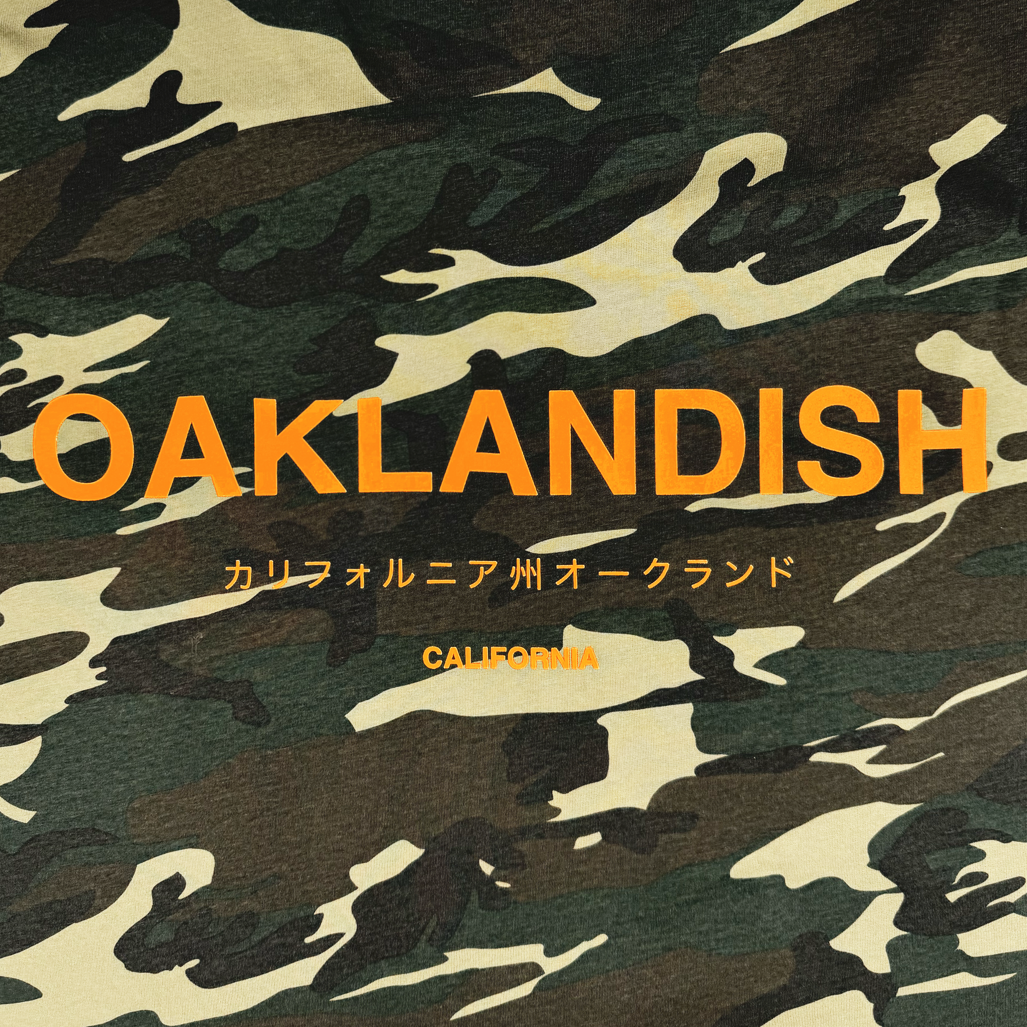 The image showcases the Modern Type Tee by Oaklandish, featuring a classic fit and camouflage design. Prominently displayed is the bold orange text "OAKLANDISH" in the center. Below are Japanese characters translating to "California Oakland," with "CALIFORNIA" in orange underneath. Crafted from 100% cotton, this tee seamlessly combines style and comfort.