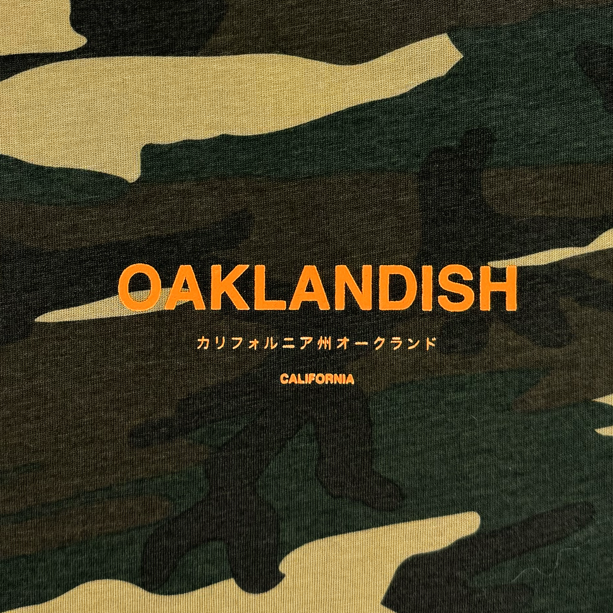 The Modern Type Tee by Oaklandish features a camouflage-patterned background with bold, orange "OAKLANDISH" text. Below, Japanese characters spell "カリフォルニア州オークランド," translating to "Oakland, California." Underneath is smaller text reading "CALIFORNIA." This classic fit t-shirt is crafted from 100% cotton with a straightforward military-inspired design.