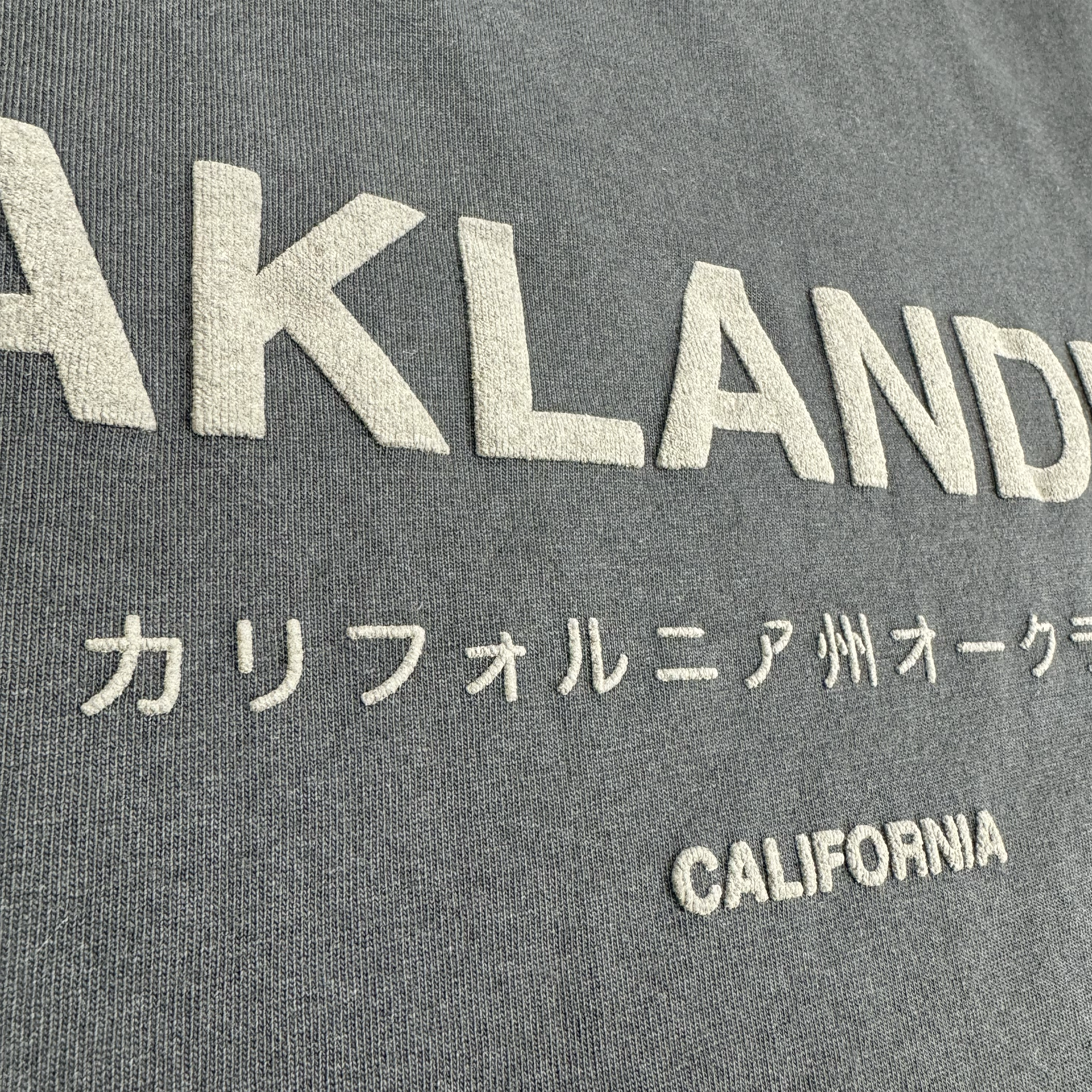 A close-up of the Modern Type Heavy Tee by Oaklandish, showcasing its heavyweight cotton fabric in a faded black color. The tee prominently features "OAKLAND" embroidered in large white letters. Beneath that, Japanese characters spell out "カリフォルニア州オークランド" (California Oakland), with "CALIFORNIA" inscribed in smaller white letters at the bottom. The texture of the fabric is clearly visible.