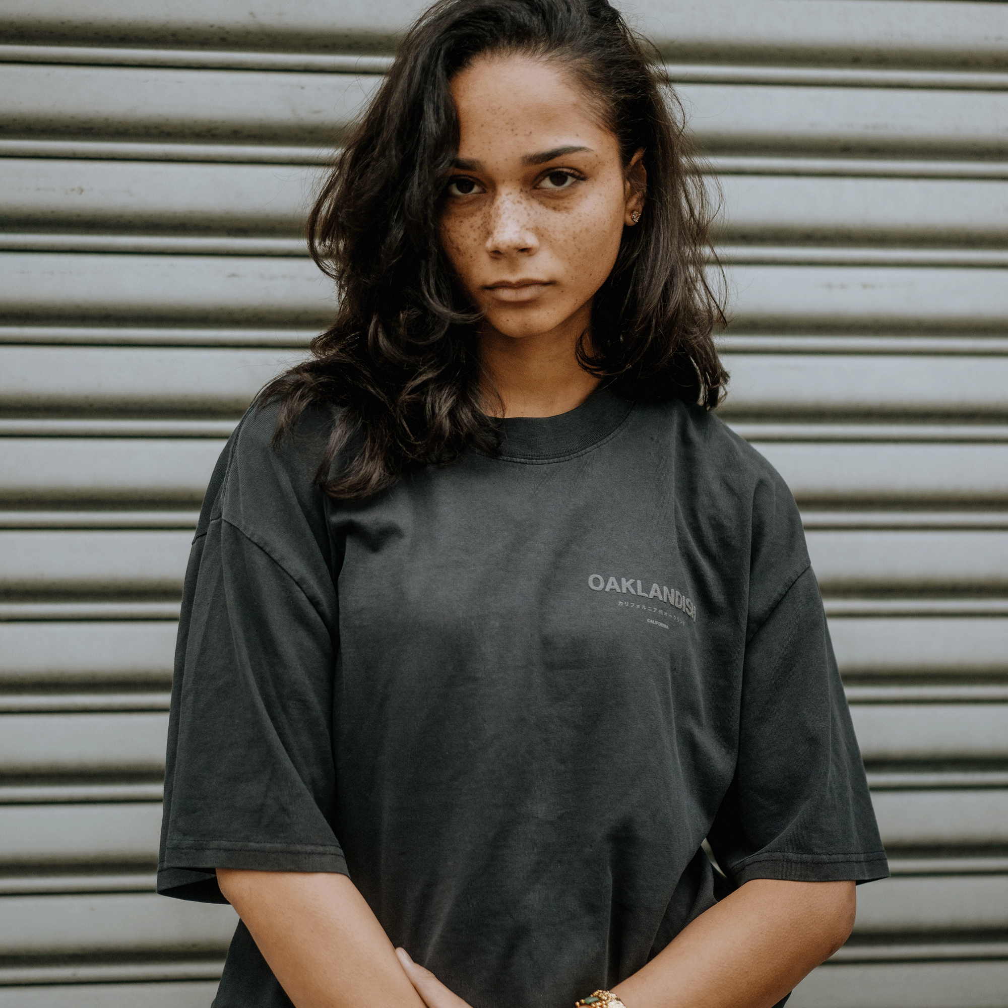 A person with long dark hair and a serious expression stands in front of a gray metal garage door. They're wearing the Modern Type Heavy Tee by Oaklandish, a faded black short-sleeved T-shirt with "OAKLANDERS" in light text on the front, featuring a boxy fit made from heavyweight cotton.