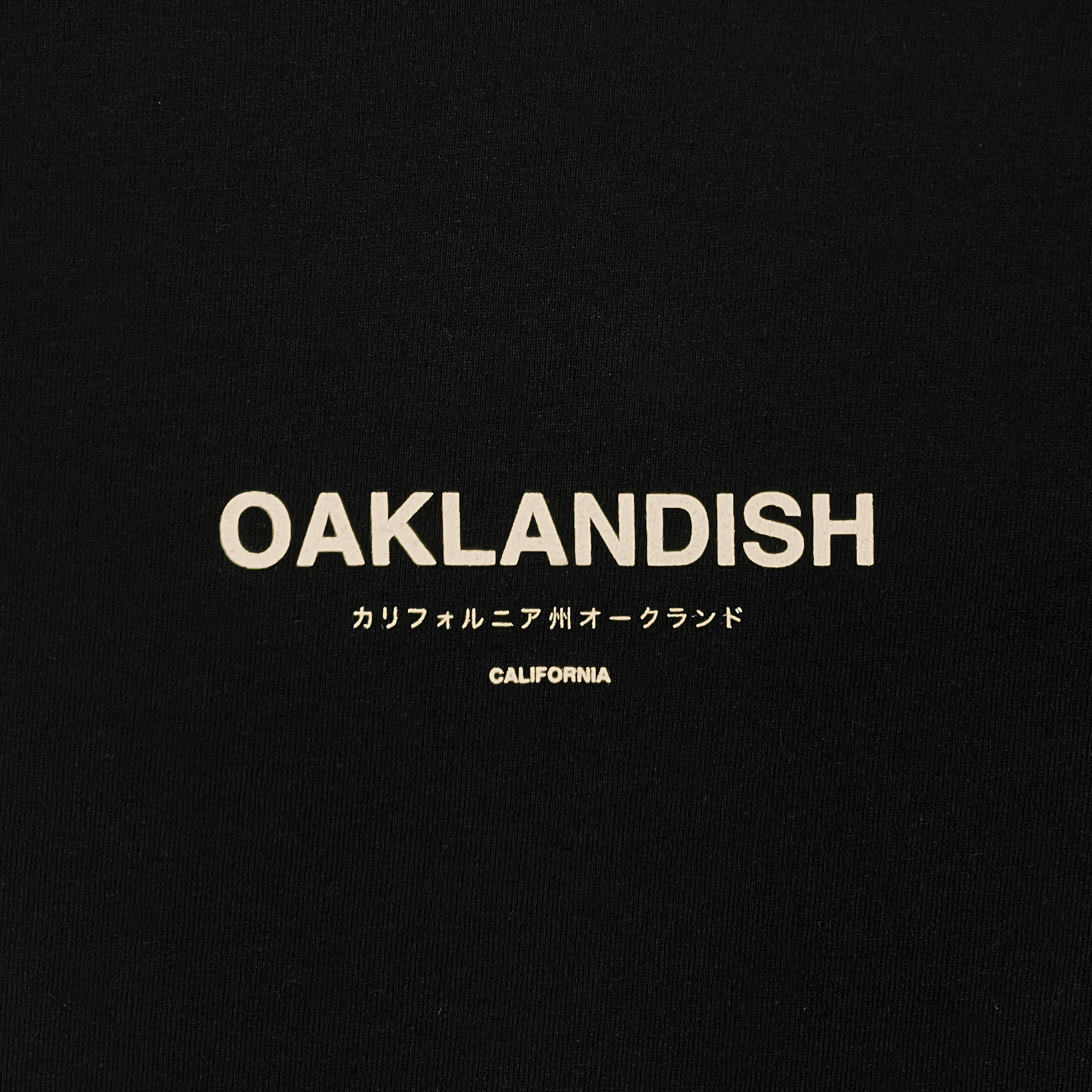 A pair of sweatpants with a black background features the word "OAKLANDISH" in bold, white, all-caps text. Below this are smaller Japanese characters, and further down, the word "CALIFORNIA" is displayed in a smaller all-caps font. These unisex Modern Type Sweatpants by Oaklandish are crafted from organic cotton.