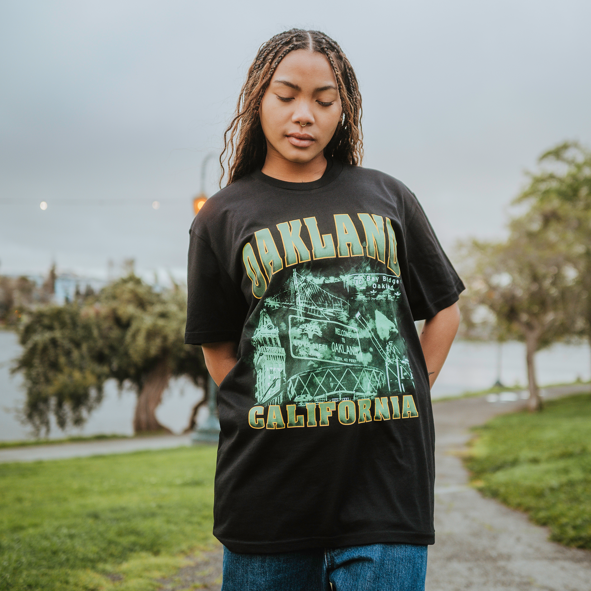 On a grassy path, a person with braided hair stands outdoors, wearing an Oaklandish Montage Tee featuring "OAKLAND CALIFORNIA" in bold green and city graphics. Their hands are behind their back and eyes closed, capturing Emerald City vibes under a cloudy sky surrounded by trees.
