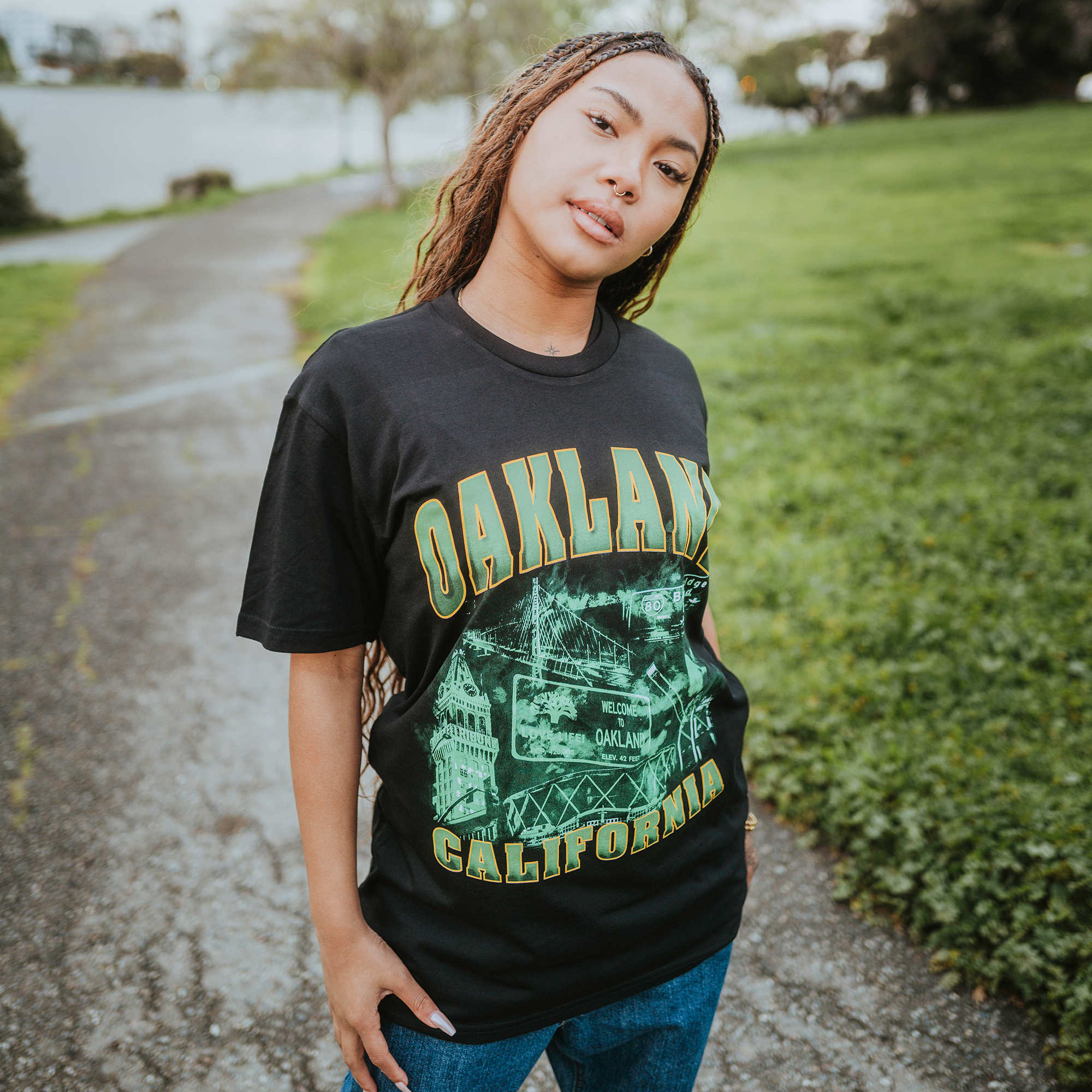 Confidently standing on a park path, they wear an "Oaklandish" Montage Tee capturing the city's essence paired with classic blue jeans. Their long braided hair flows as they look into the camera, surrounded by lush trees and grass under an overcast sky.