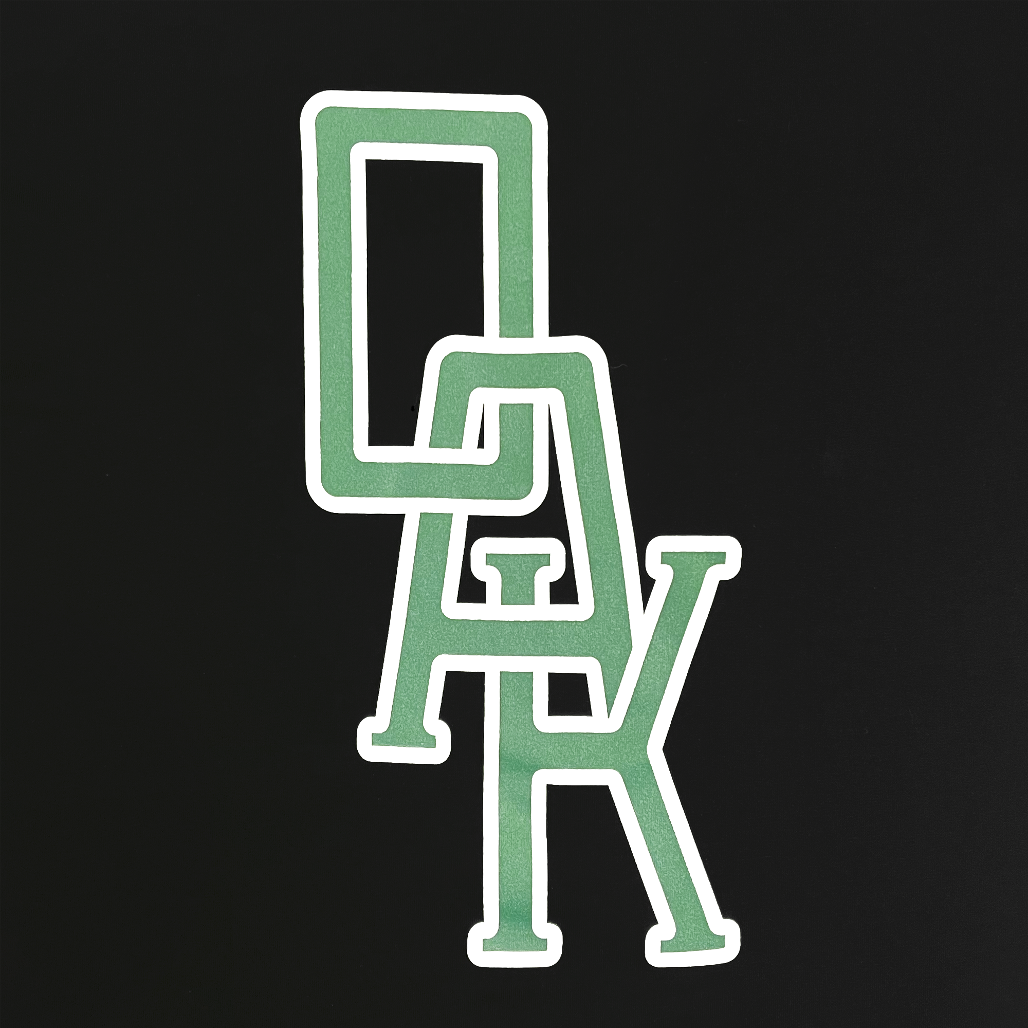 The image showcases the Oaklandish OAK Monogram Tee, featuring vertically interlinked uppercase "O," "A," and "K" letters, outlined in white and filled with green against a black background. The classic fit t-shirt is made of 100% cotton, with the "O" at the top, "A" in the middle, and "K" at the bottom.