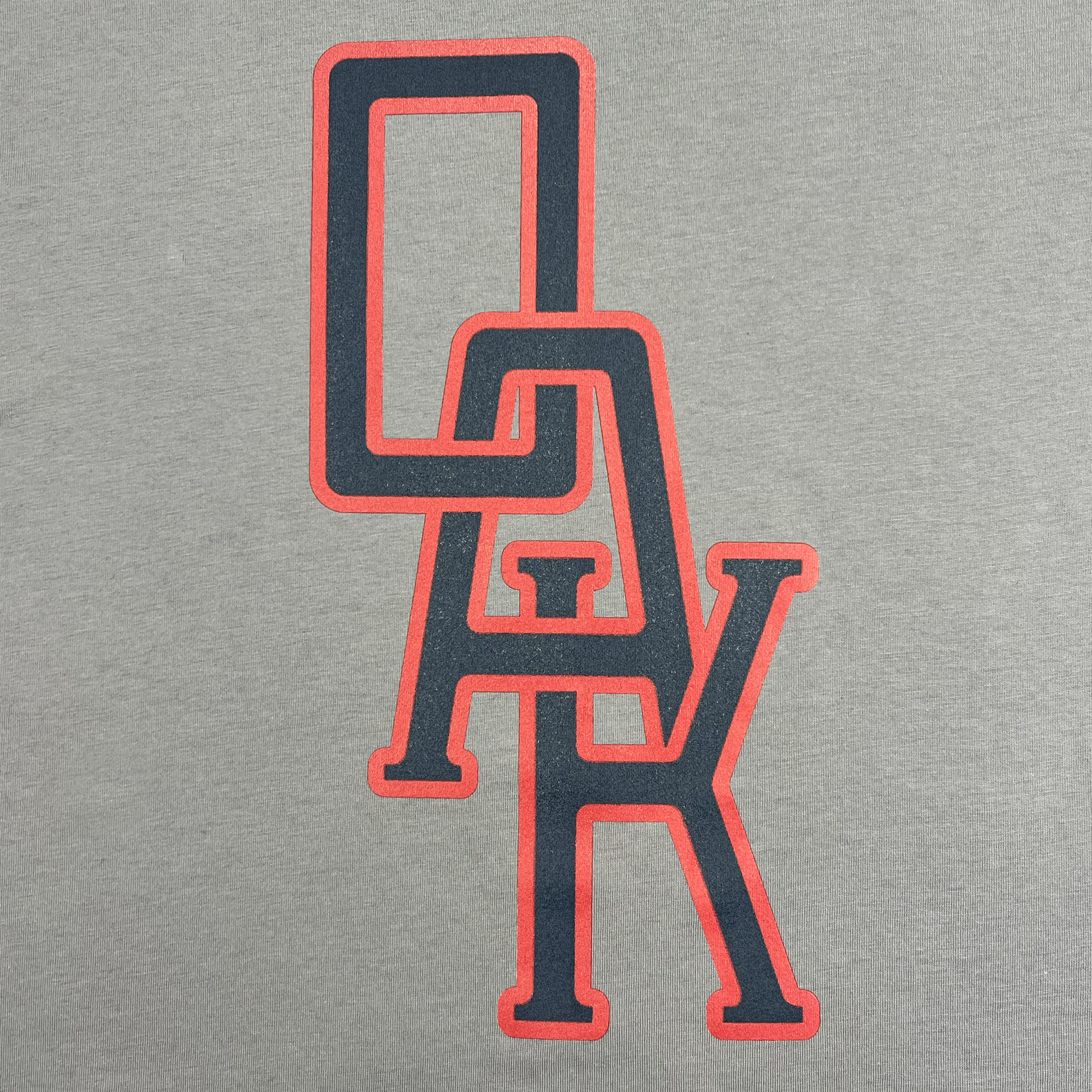The OAK Monogram Tee by Oaklandish is adorned with a stylized graphic featuring the letters "OAK" in a bold, overlapping design. The large "O" is red with a black interior, placed over a black "A" with red edges, and the "K" is black with a red outline. This classic fit t-shirt is crafted from 100% cotton and features the design centered on a granite grey background.