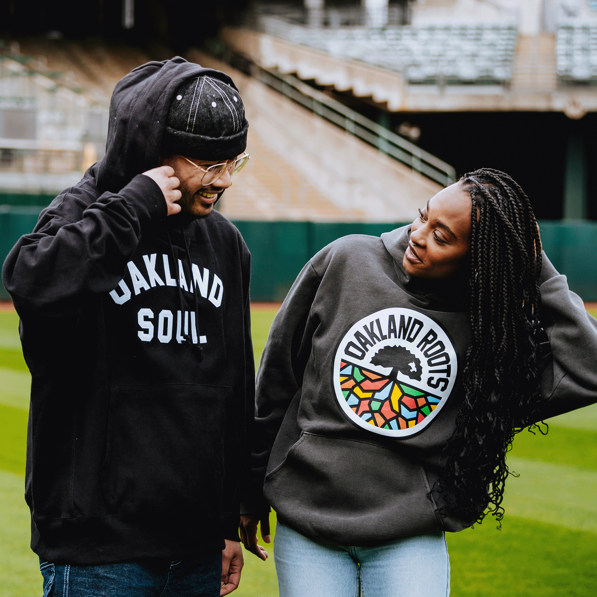 Two people smile at each other on a baseball field. One wears an Oaklandish “OAKLAND SOUL” hoodie, while the other sports an Oakland Roots SC Heavy Hoodie by Oakland Roots SC—each proudly repping their favorite local club.