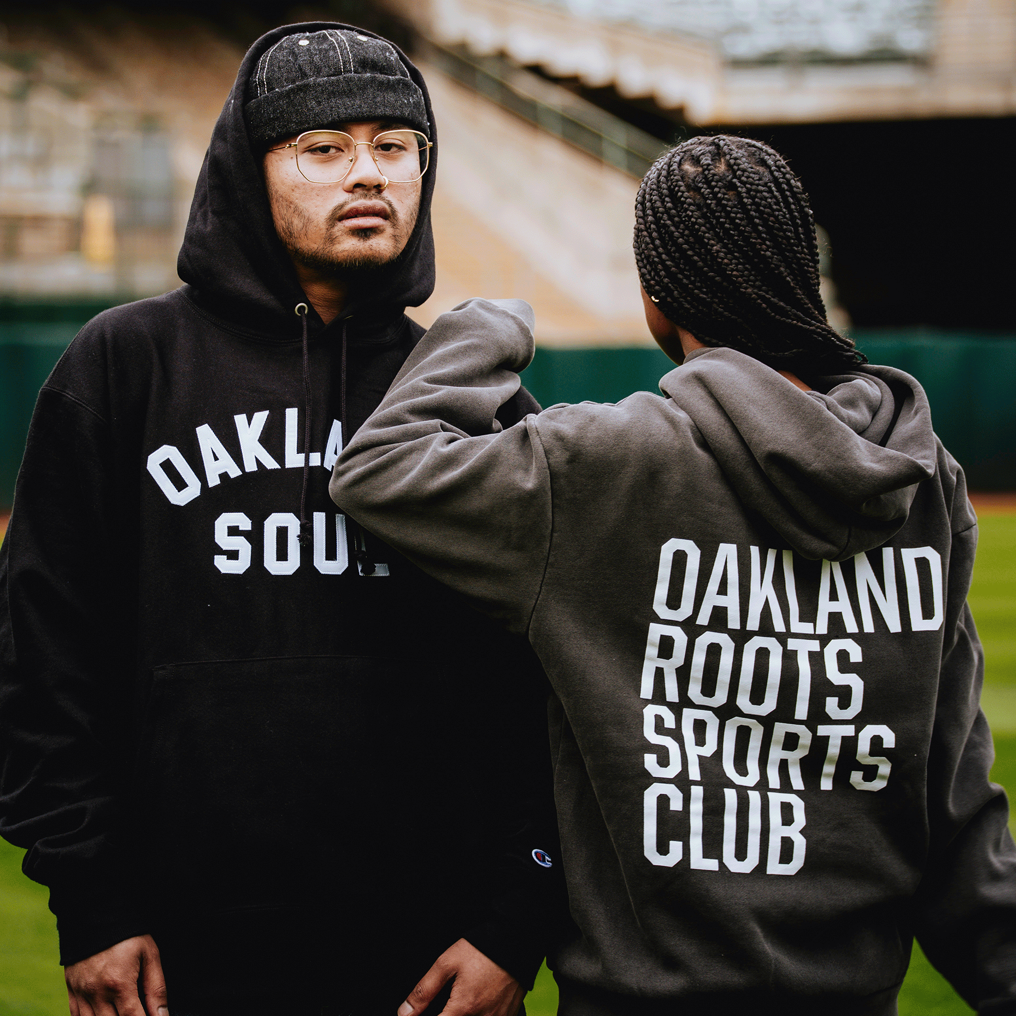 Two people stand on a sports field in hoodies. One, facing forward in glasses, wears a black Oaklandish Pullover Hoodie; the other, with braided hair and back turned, is in an Oakland Roots SC Heavy Hoodie. Stadium seating appears blurred behind them.