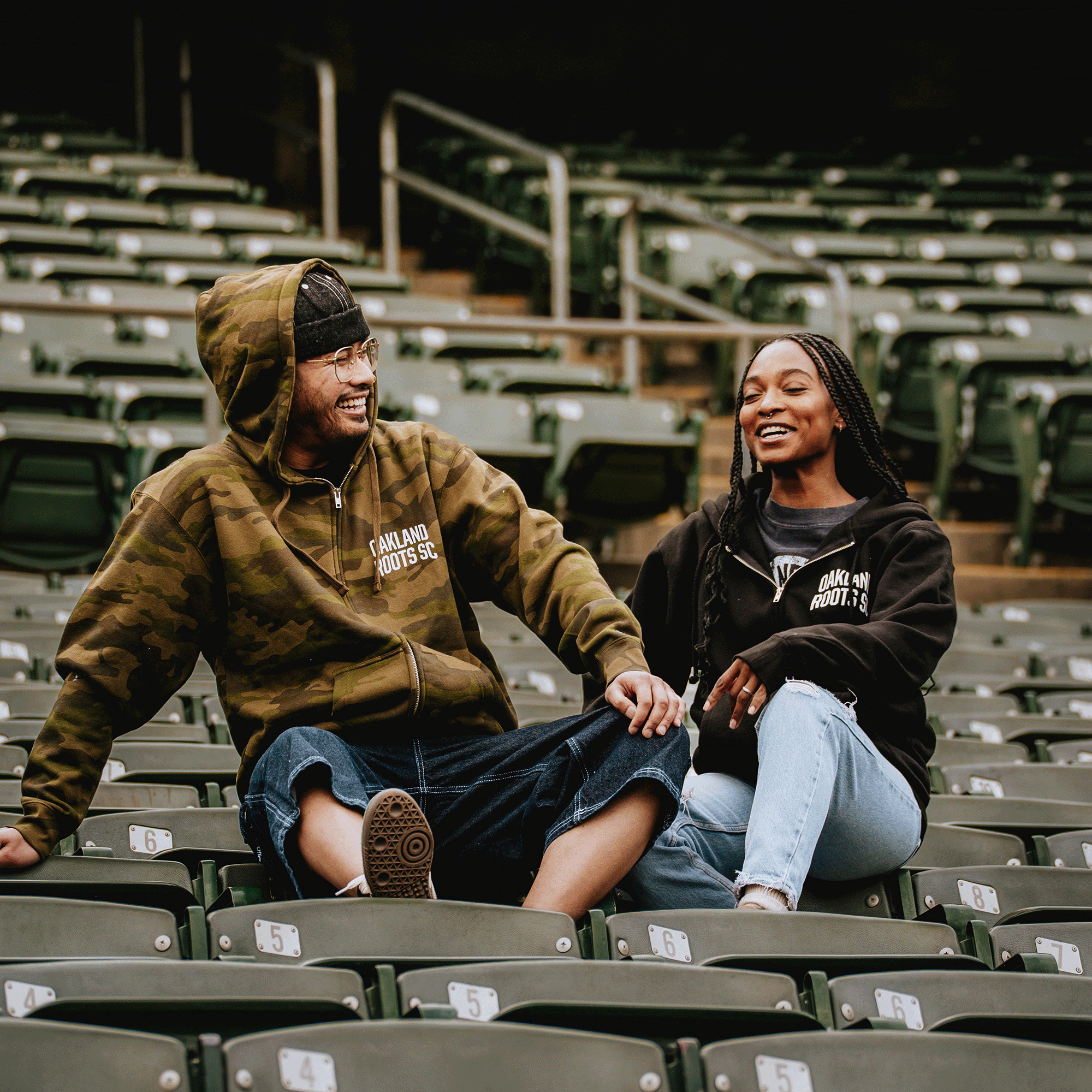 A man and woman smile on stadium seats wearing Oakland Roots SC Classic Zip Hoodies, showing support for the brand’s commitment to social good. They sit relaxed and happy together among mostly empty green seats.