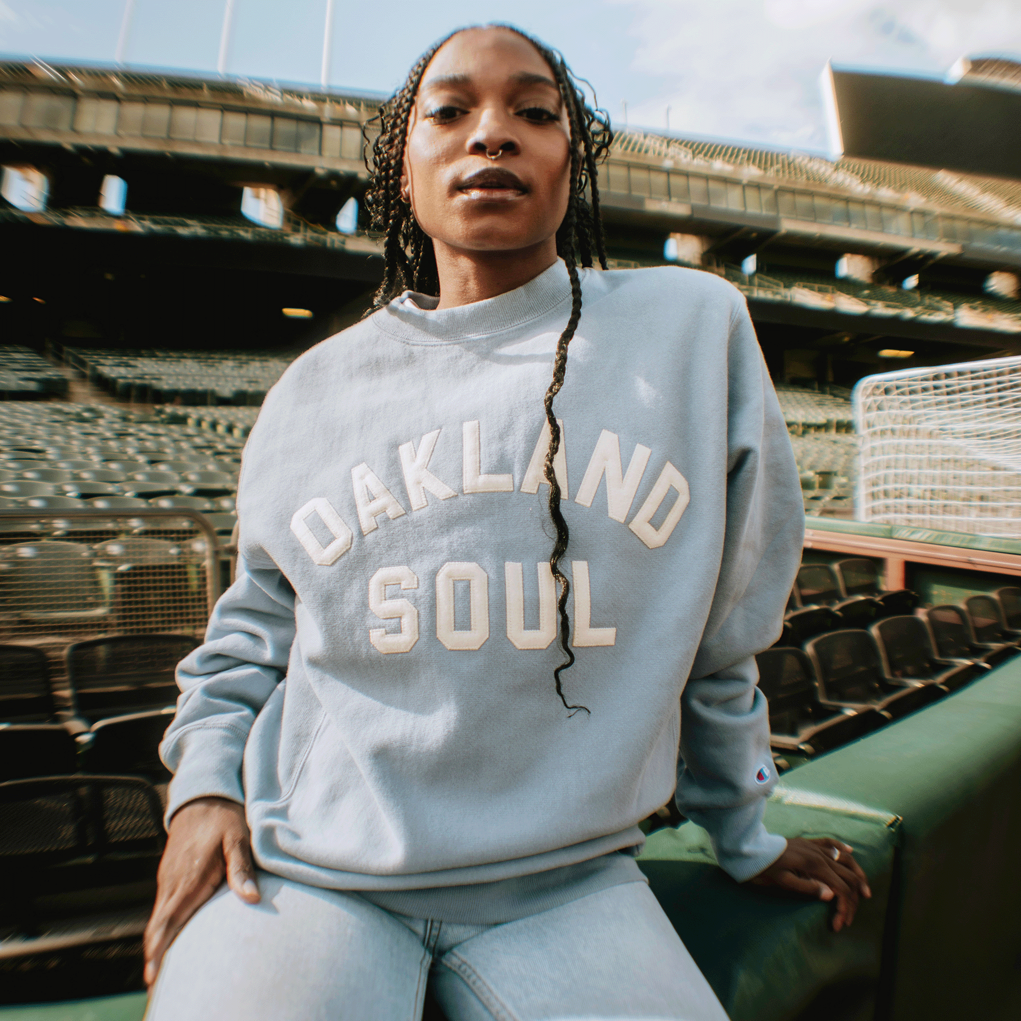 A woman with braided hair sits in stadium seats, confidently wearing the Oakland Soul Champion X Oakland Soul Wordmark Crew sweatshirt. The empty stands highlight women’s soccer and its positive social impact under a partly cloudy sky.