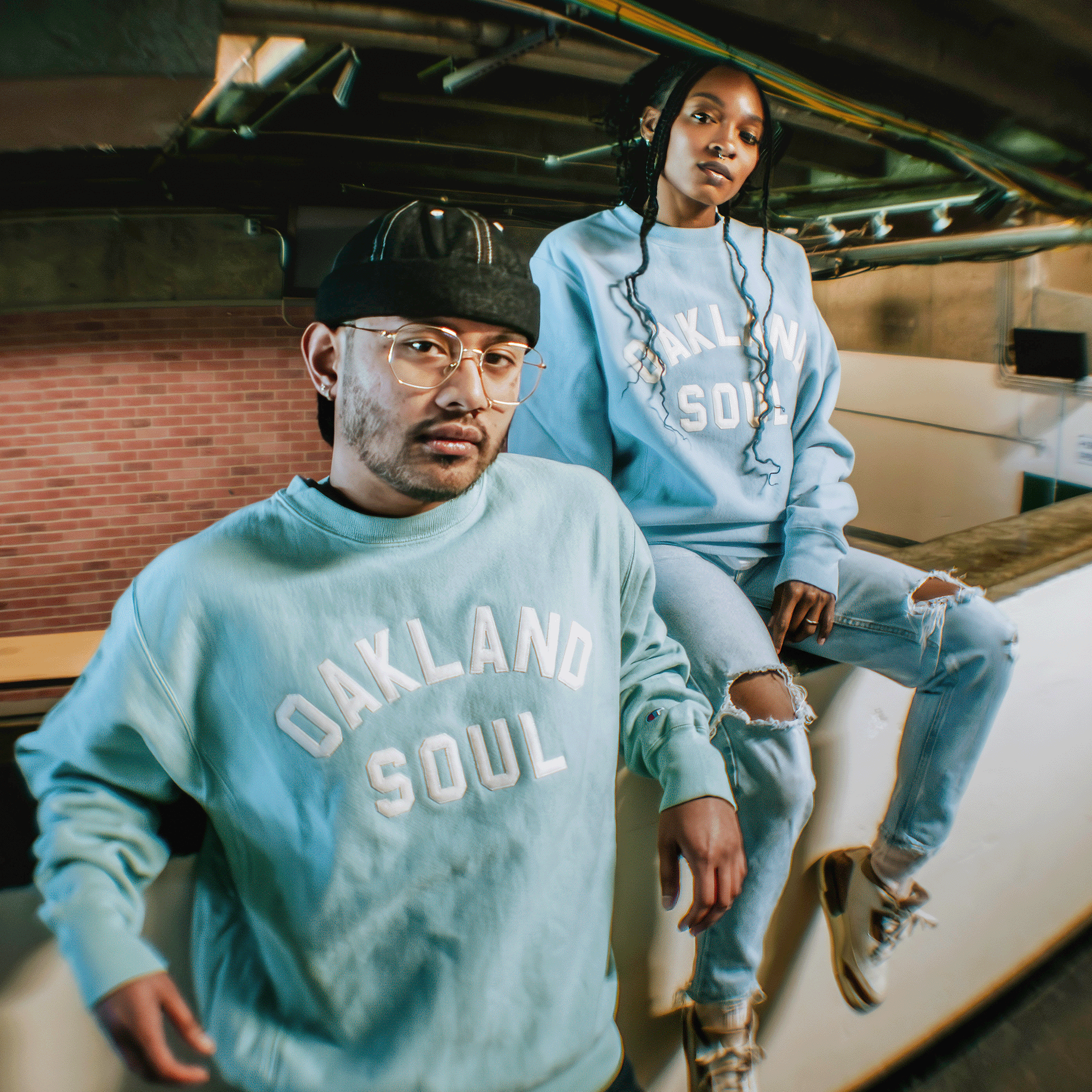 Two people showcase Oakland Soul’s Champion X Oakland Soul Wordmark Crew indoors—one sitting, one standing—against a brick and industrial backdrop, both confidently representing the women’s soccer brand.
