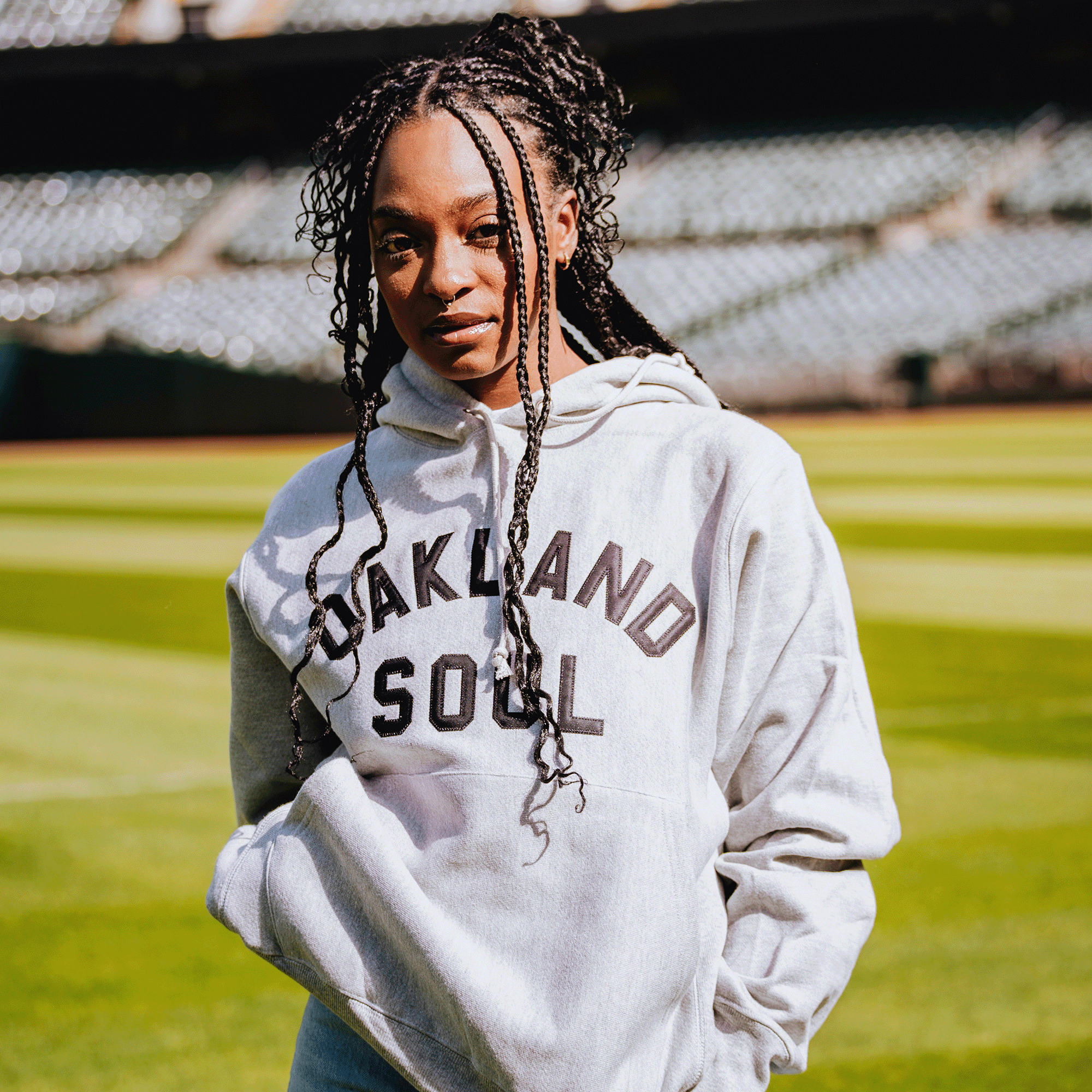 A young woman with braided hair stands on a well-lit sports field, confidently wearing the Champion X Oakland Soul Wordmark Hoodie by Oakland Soul, the USL W-League women’s soccer team. Blurred stadium seats fill the background.