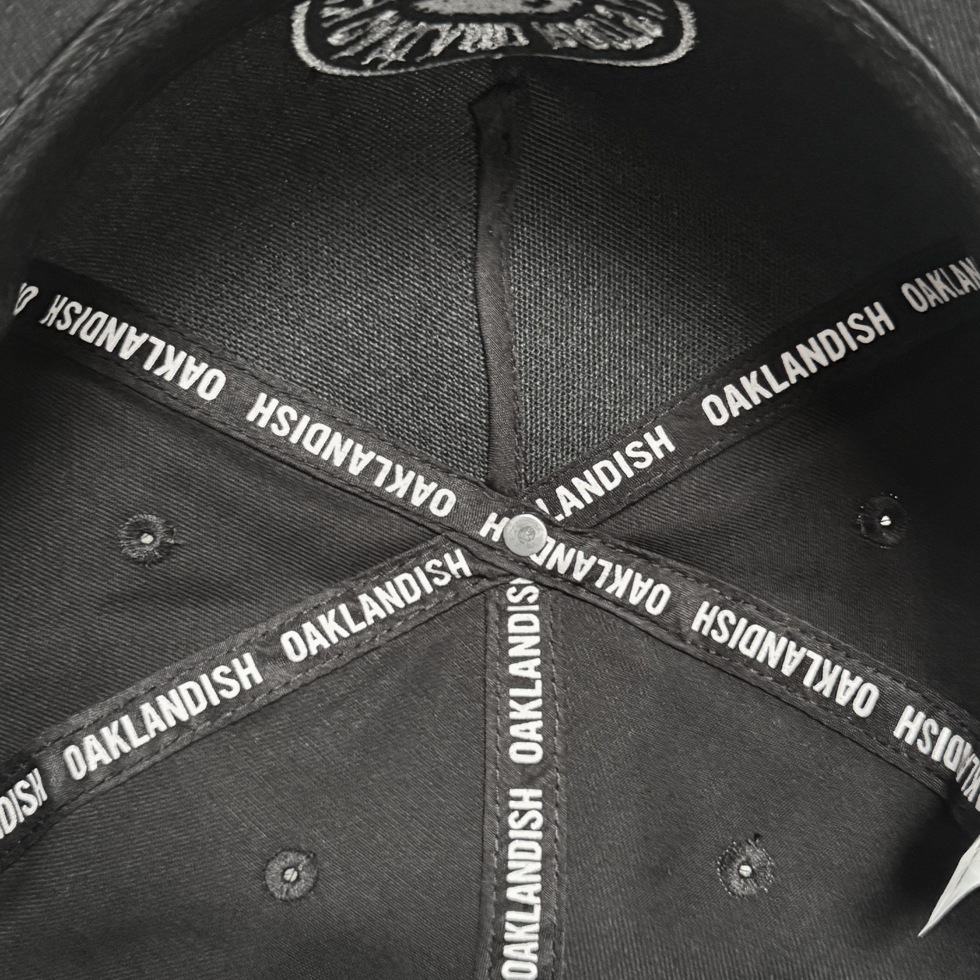 Inside view of the ORSC 5 Panel Hat by Oakland Roots SC, a top Oakland sports item. Intersecting fabric strips labeled “OAKLANDISH” form a star around a central button, with visible stitching and metal eyelets on black fabric.