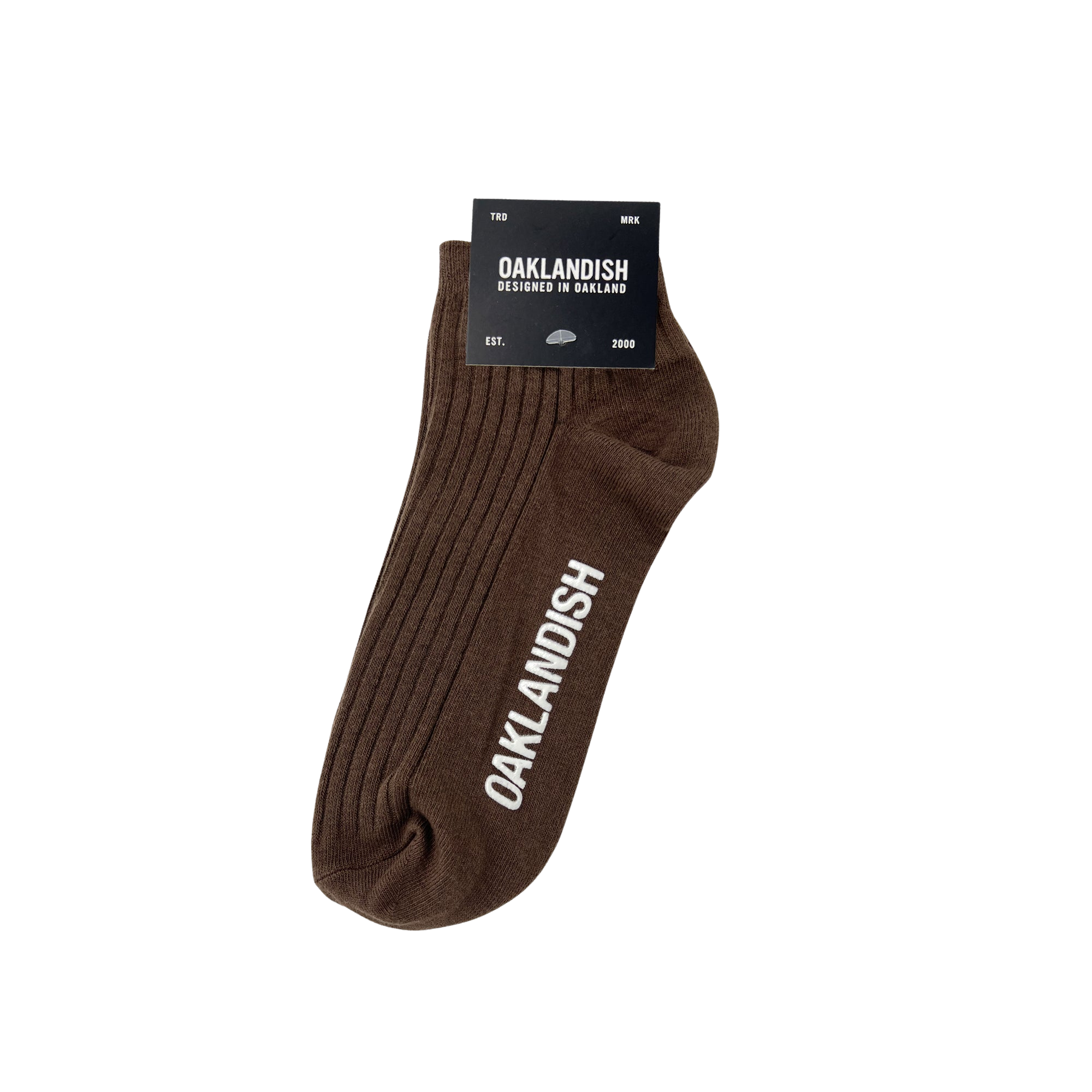 A single brown Oaklandish Grip Low Crew Sock displayed against a white background. The sock features vertical ribbing and the word "OAKLANDISH" in white capital letters along the side. Attached to the sock is a black label with white text detailing the brand name "OAKLANDISH" and other product information.