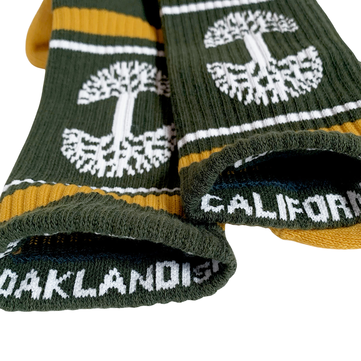 Close-up of two forest green and yellow OAK Knit Crew Socks from Oaklandish, featuring white trees and stripes near the cuffs. The word "CALIFORNIA" is visible on one sock's foot area, and "OAKLANDISH" on the other. These crew socks are displayed on a white surface and are made from cotton, polyester, and spandex.