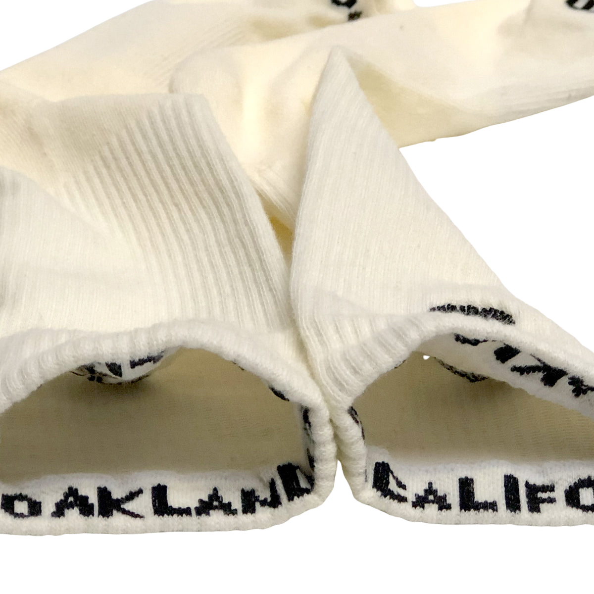 Two off-white cotton crew socks from Oaklandish, known as Embroidered Logo Socks, are laid out with their cuffs turned inside out. The cuffs have a ribbed texture and feature the text "OAKLAND" in black capital letters at the interior edge. These socks are positioned so that the text is partially visible.