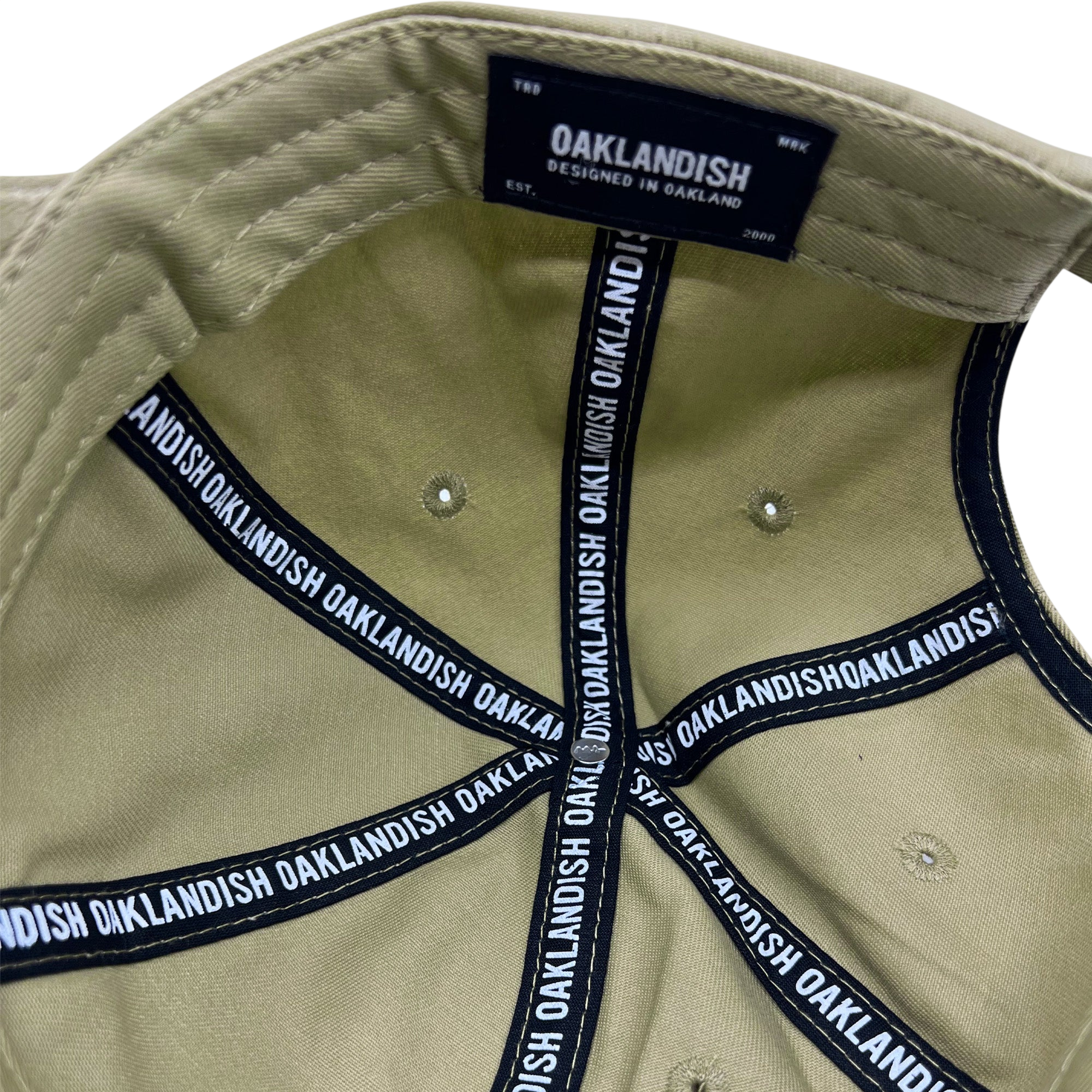 The image reveals the interior of an Oaklandish Official Micro Logo Dad Hat. The black fabric bands, imprinted with "Oaklandish" in white, fan out from the center of the crown. Inside, a tag reads, "Oaklandish, designed in Oakland, Est. 2000." The hat features an adjustable strap for a perfect fit.