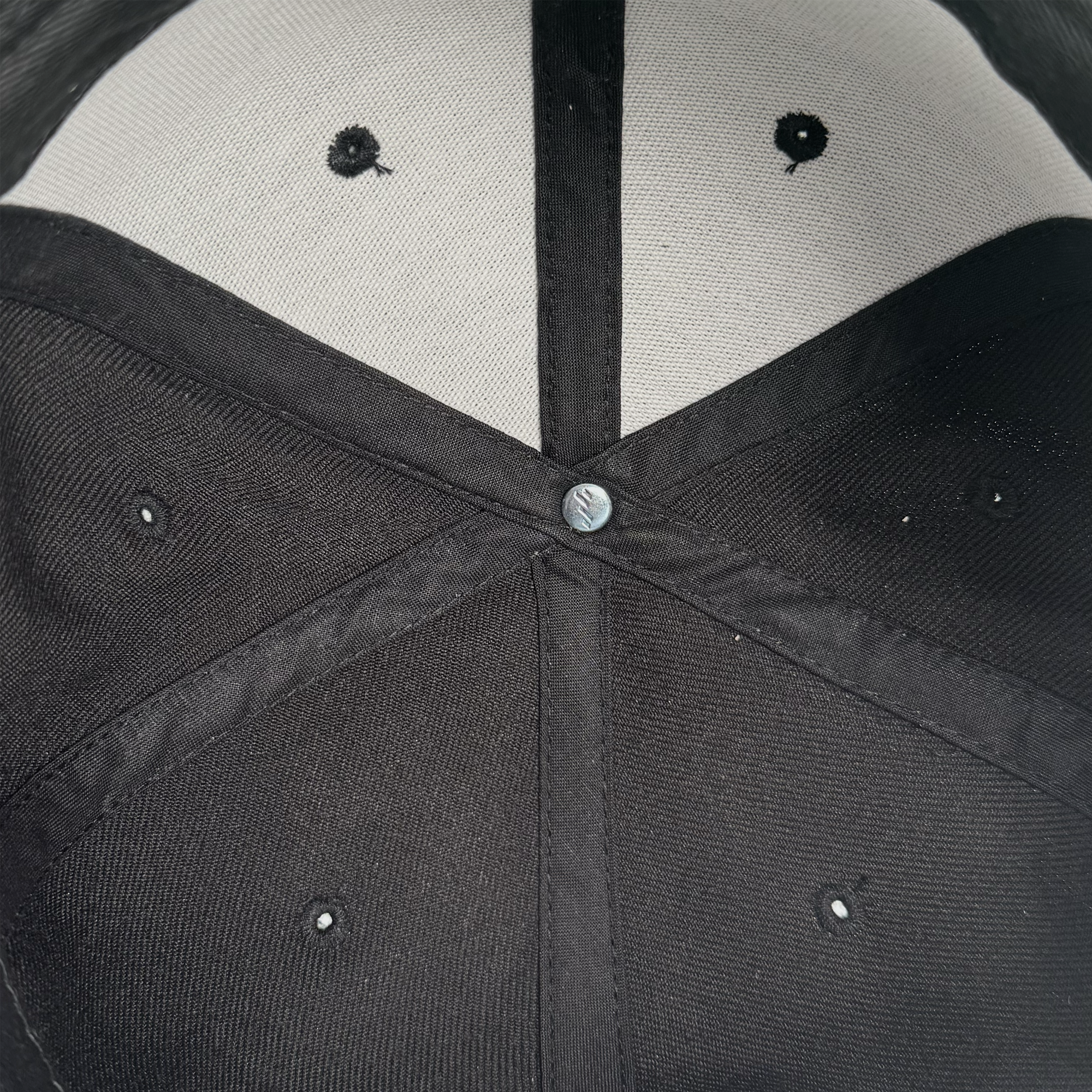 Close-up of the inside top of the black "Power of the People Snapback" by Oaklandish, showing stitched panels, a silver button at center, small ventilation holes, white fabric lining, and stitching forming an "X" around the button.