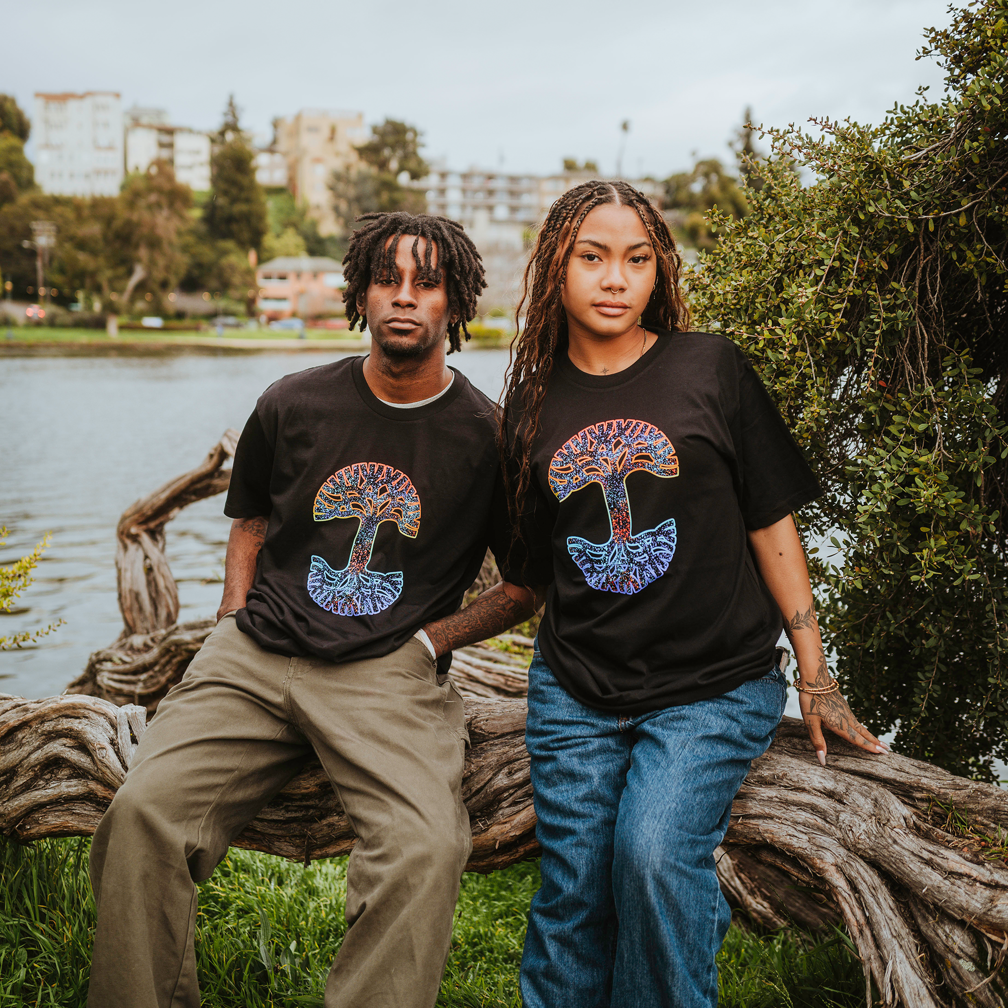 A man and woman sit on a twisted tree trunk by the lake wearing Oaklandish Prism Tees with colorful mushroom designs. She’s in braids and jeans, he has short locs and corduroy pants. Tall trees and buildings loom under the cloudy sky.