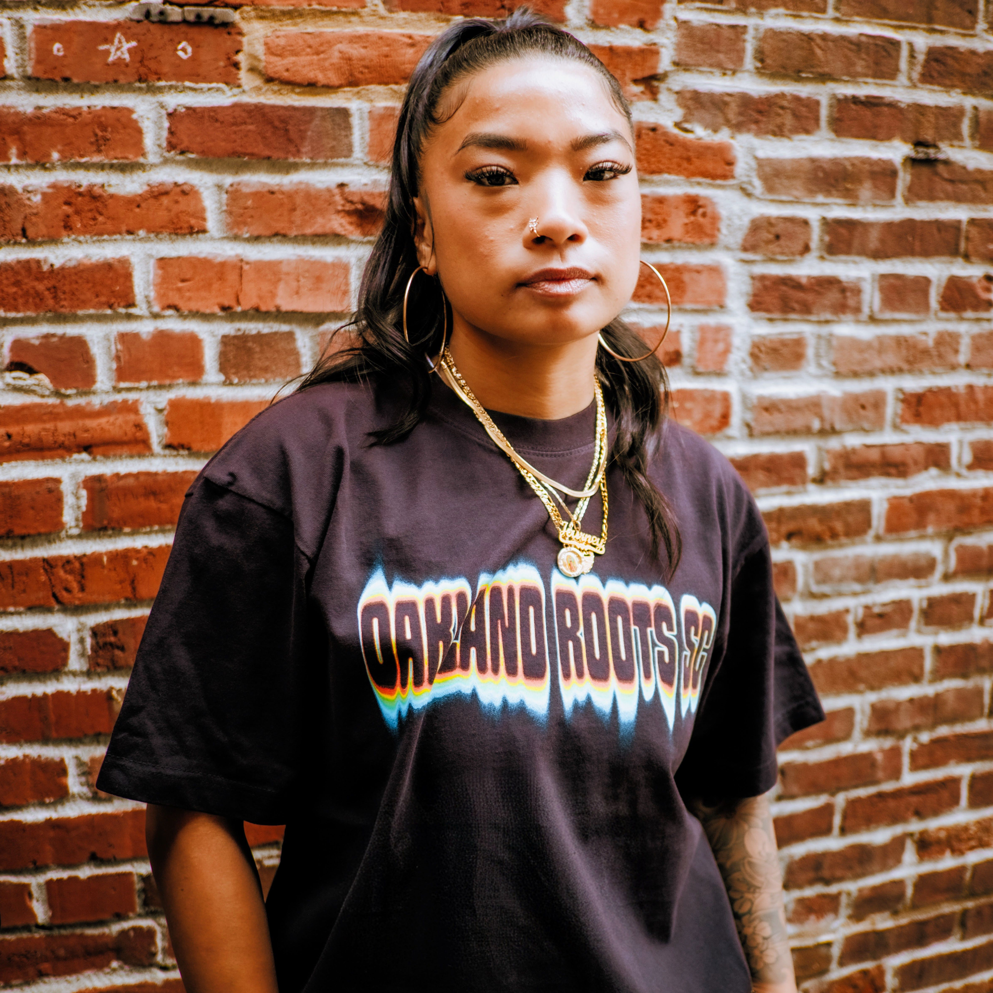 A person with long dark hair in a high ponytail stands against a brick wall, wearing gold hoop earrings, layered gold necklaces, and a black Oakland Roots SC Acid Tee. They look confidently at the camera with a calm expression.