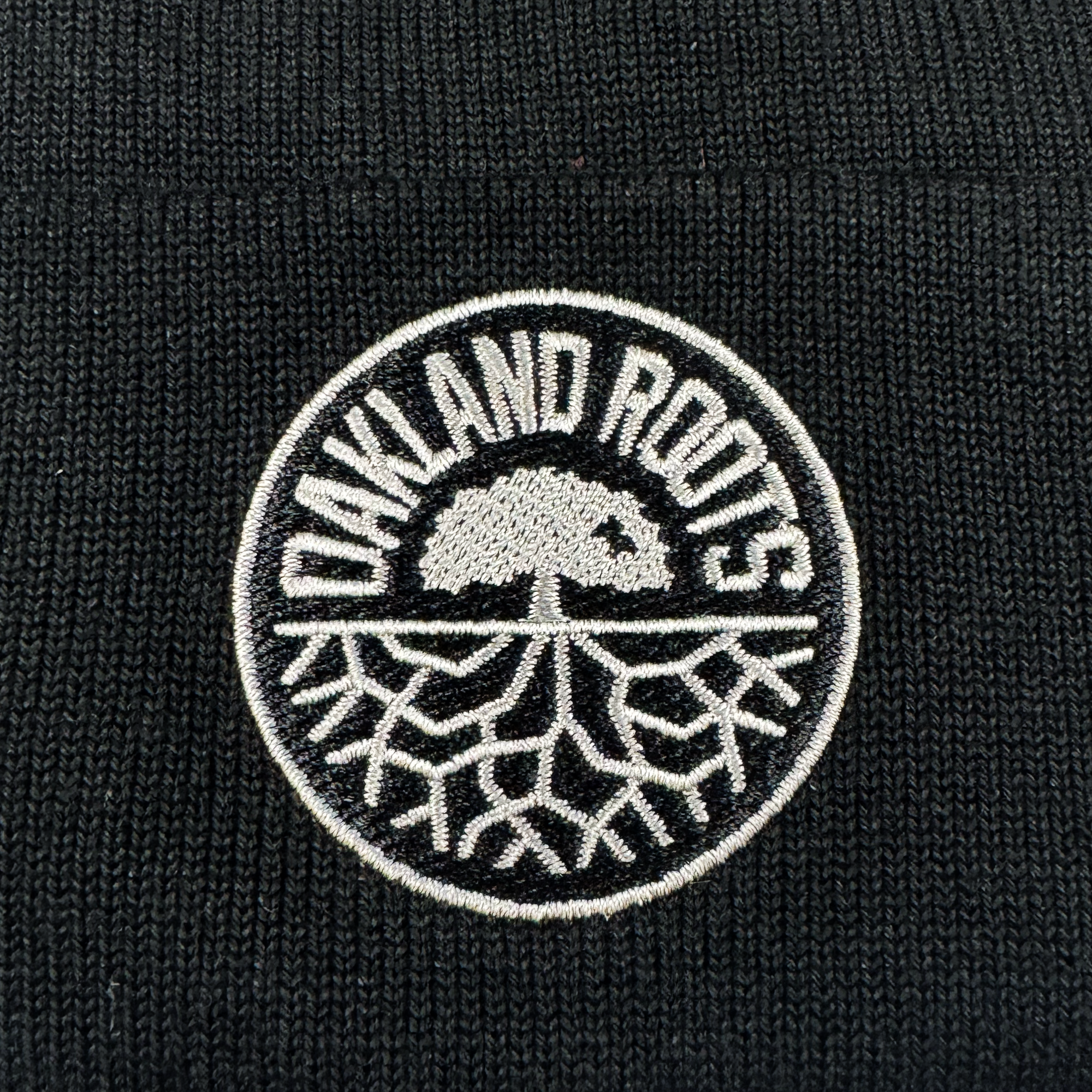 A close-up of the black ORSC Beanie by Oakland Roots SC, showcasing a circular white-embroidered patch with “OAKLAND ROOTS” and a solid-canopy tree with exposed roots.