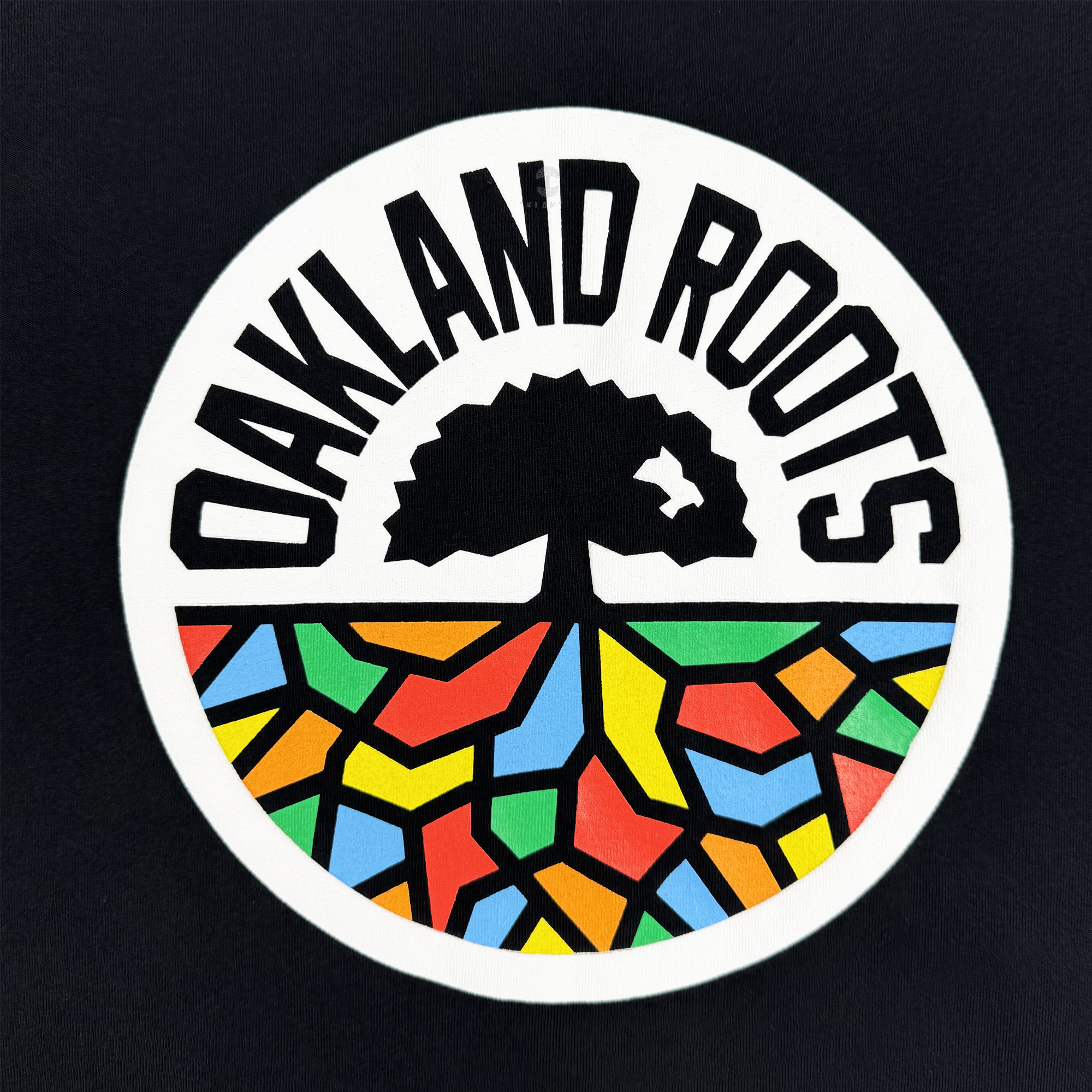 The Oaklandish Bootleg Tee features a circular “OAKLAND ROOTS” logo above a black tree with colorful mosaic roots on white, inspired by Oaklandish style—all set against a bold black backdrop for the Oakland Roots SC.