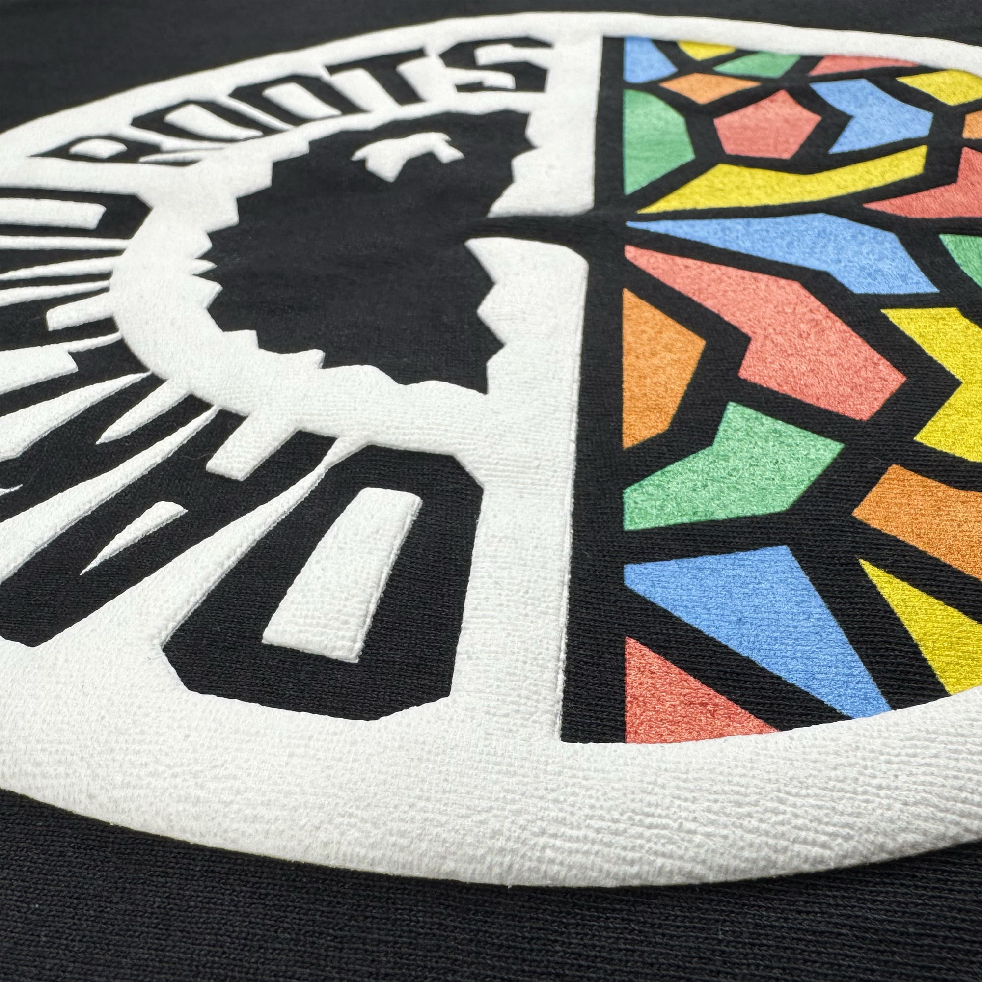 Close-up of textured black fabric on the Oakland Roots SC X Oaklandish Bootleg Tee by Oaklandish, featuring raised embroidery of a circular motif blending Africa’s silhouette and stained-glass patterns, with partial “ROOTS” text.