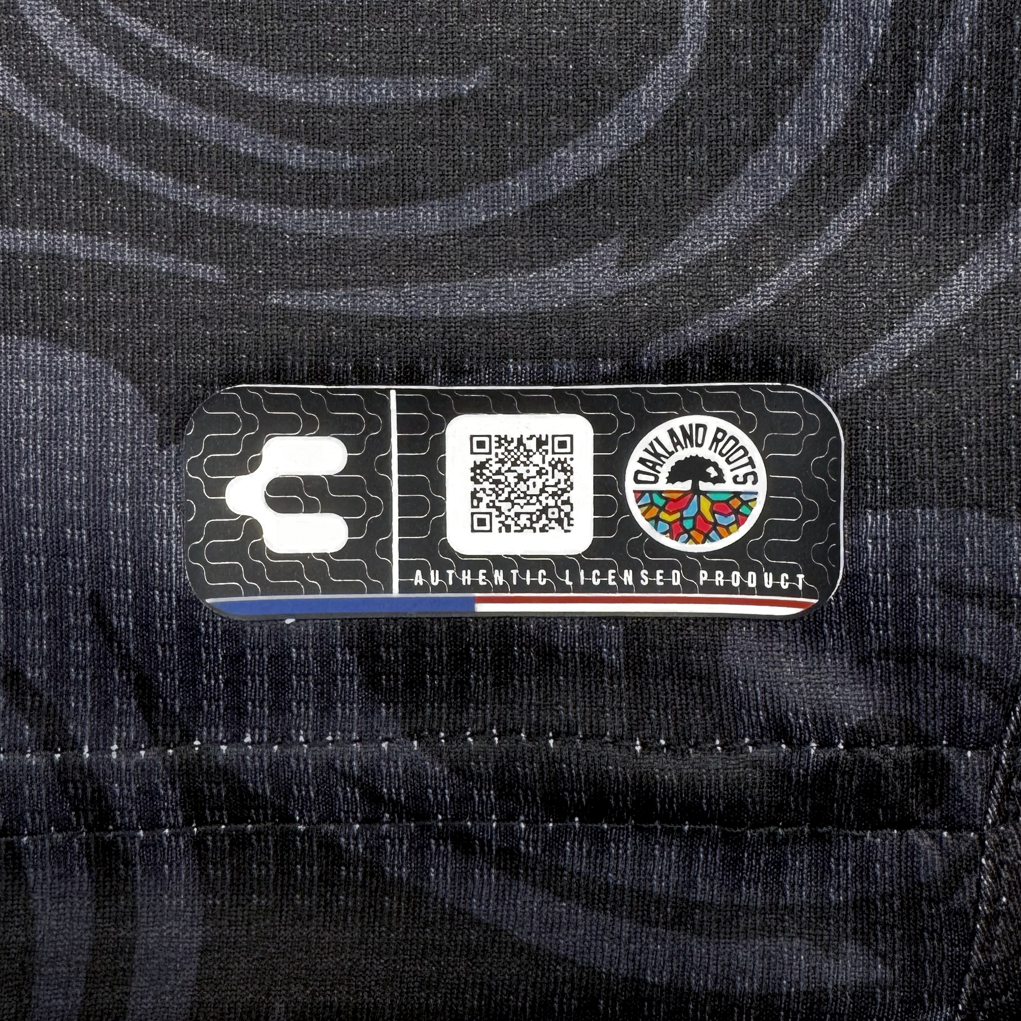 Close-up of a rectangular black patch on fabric from the Custom Unisex Oak-ness Awakens Kit by Oakland Roots SC. The patch shows a white “C,” QR code, Oakland Roots logo with multicolored roots, and “AUTHENTIC LICENSED PRODUCT.”.