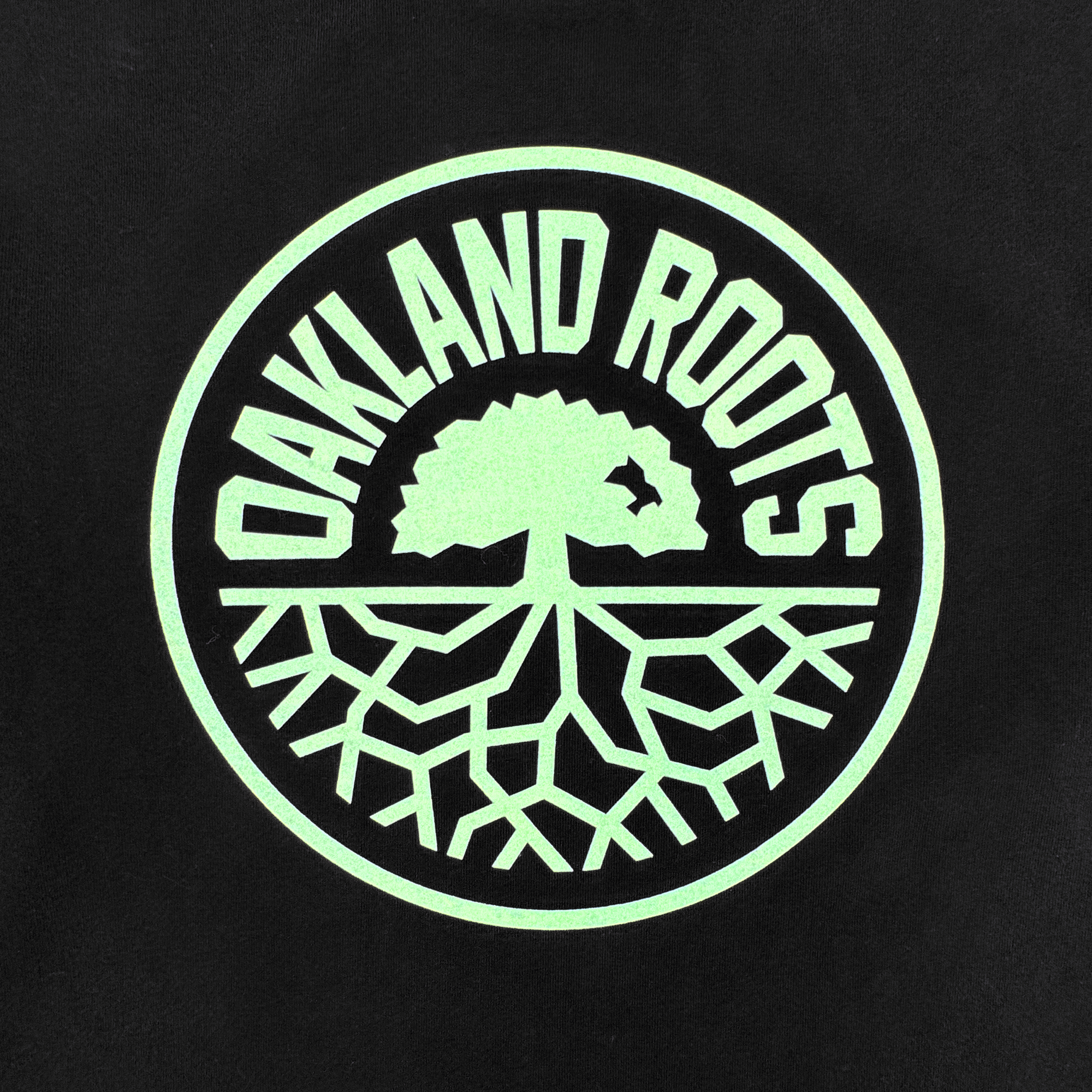 A circular Oakland Roots SC logo in light green on black features bold “OAKLAND ROOTS” text around a large tree and roots—symbolizing unity and team spirit. Perfect for any fan, this design appears on the ORSC Ryujin LS Tee.