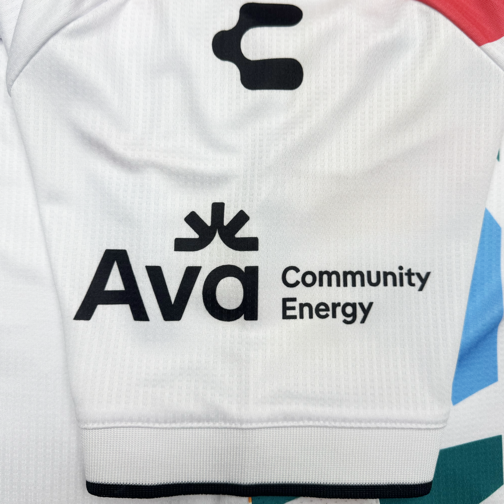 A white sleeve from the Custom Unisex Mosaic in Motion Kit by Oakland Roots SC displays a black logo and “Ava Community Energy” beneath a stylized symbol, with subtle vertical lines, a black cuff, and part of another multicolored kit visible nearby.