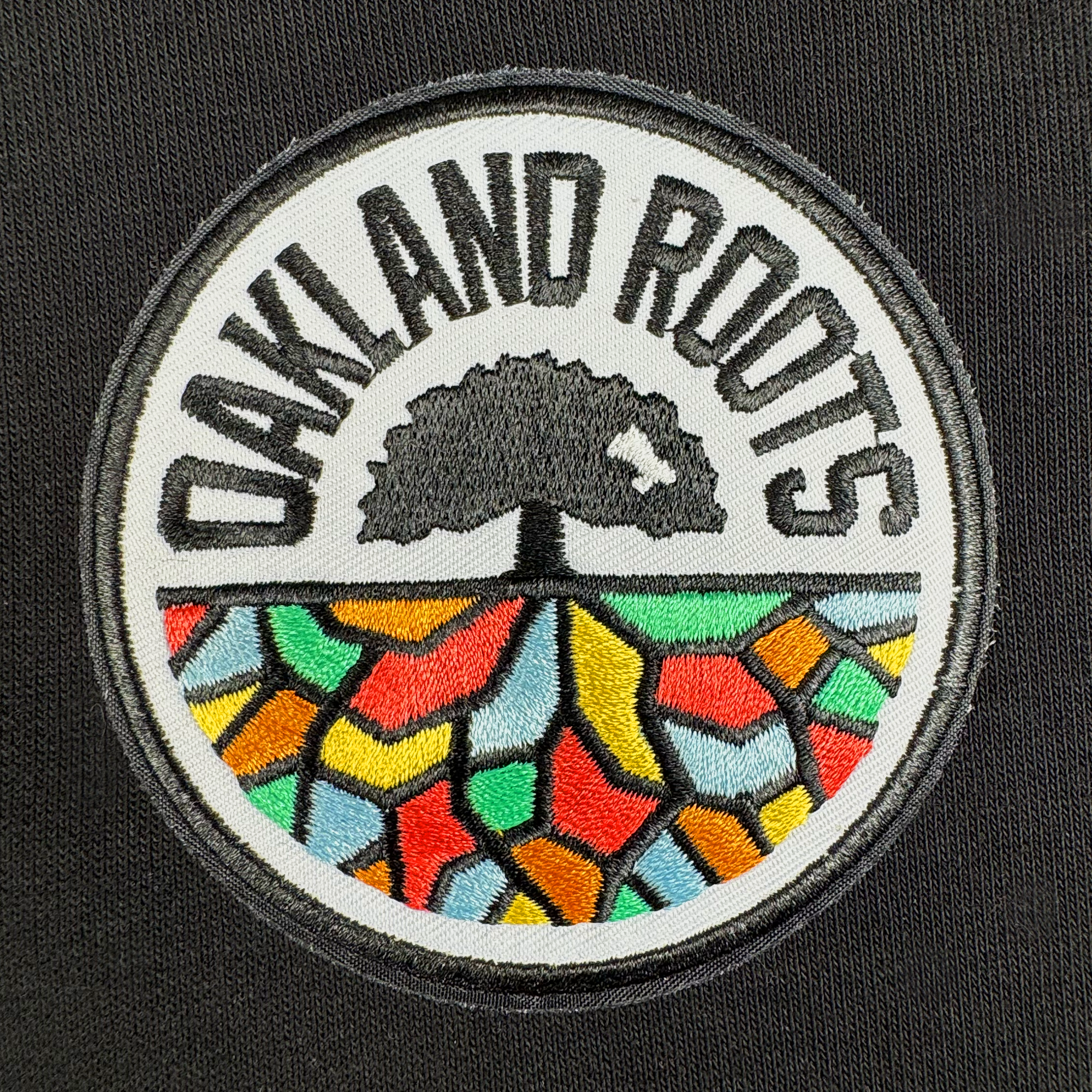 The Oakland Roots SC Logo Premium Hoodie features the club’s circular emblem with “OAKLAND ROOTS” around a black tree and colorful mosaic roots—an essential for any Oakland Roots SC fan.