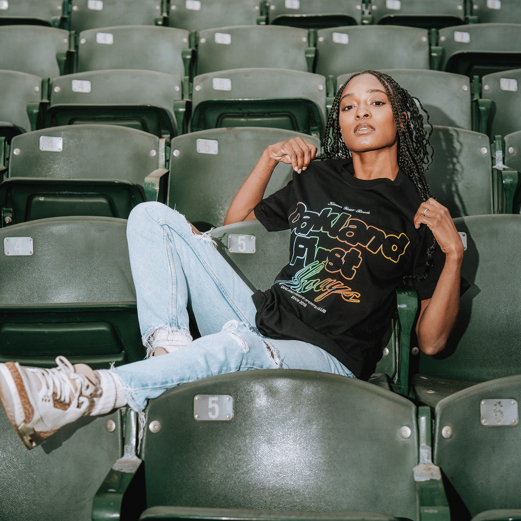 A woman with long braided hair reclines on green stadium seats, wearing light blue ripped jeans, white sneakers, and the Oakland Roots SC OFA Different Tee. She looks confidently at the camera, showing pride in Oakland Roots.