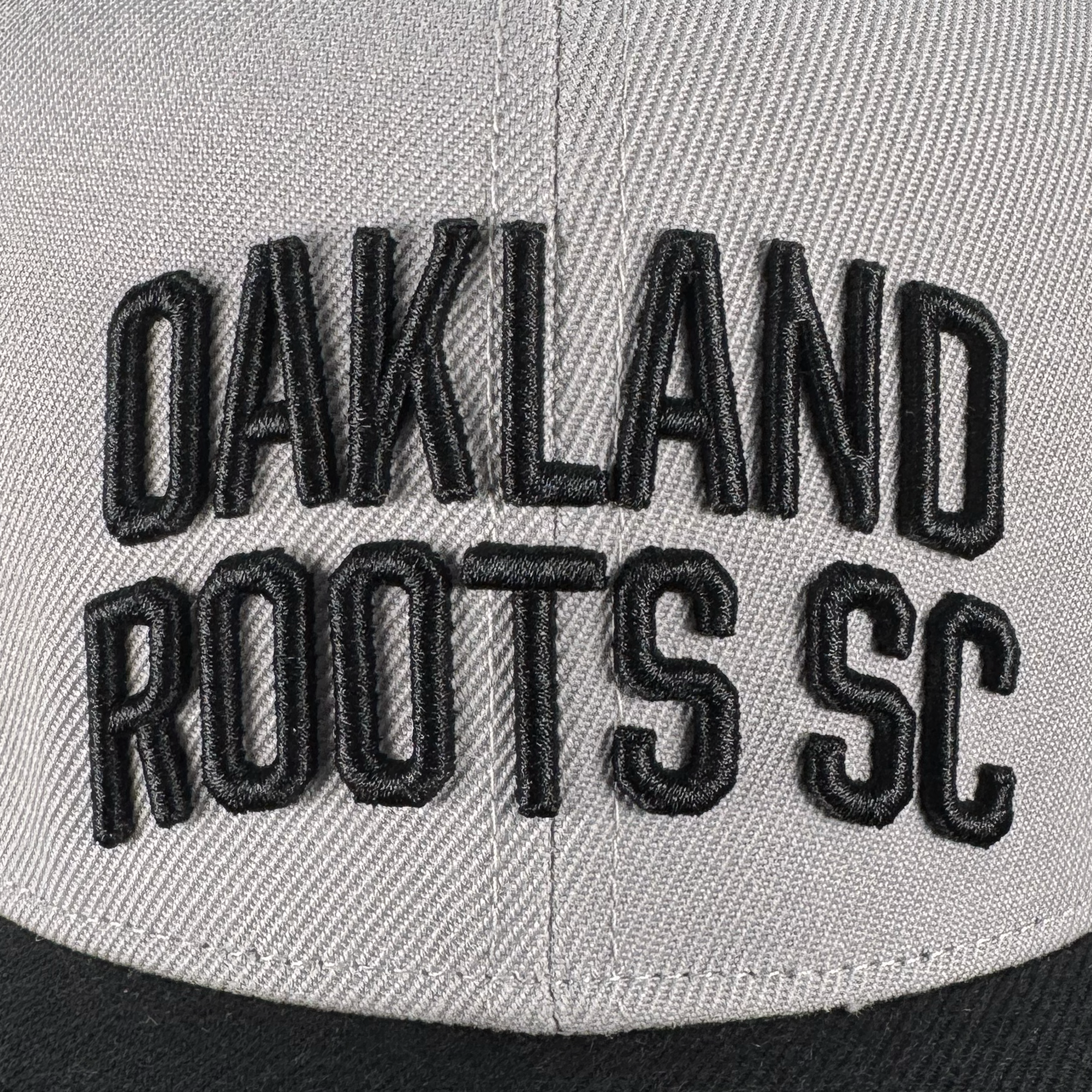 Close-up of the Oakland Roots SC Arch Text Snapback by Oakland Roots SC, featuring bold black "OAKLAND ROOTS SC" embroidery on light gray fabric and a partially visible black bill. Raised stitching enhances the arch text design.
