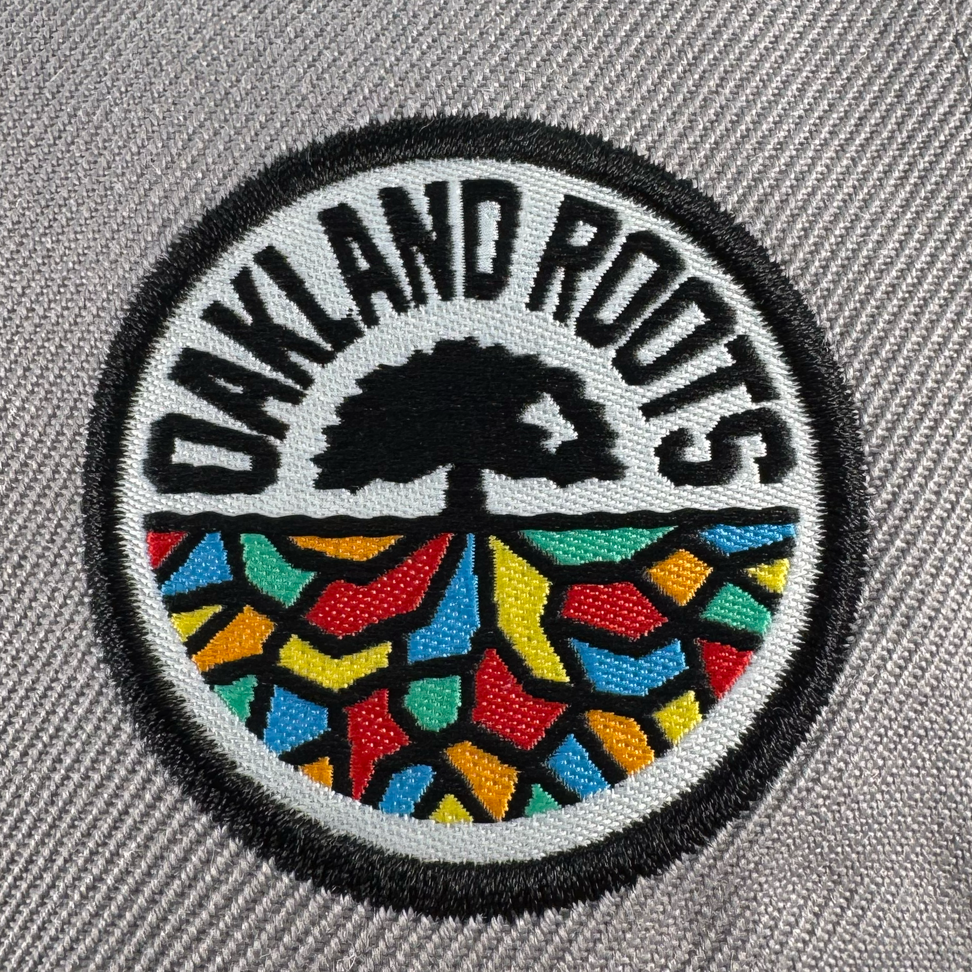 The Oakland Roots SC Arch Text Snapback features a circular embroidered patch with bold “OAKLAND ROOTS,” a black tree, and vibrant mosaic ground. The patch is sewn onto a stylish gray textured snapback from Oakland Roots SC.
