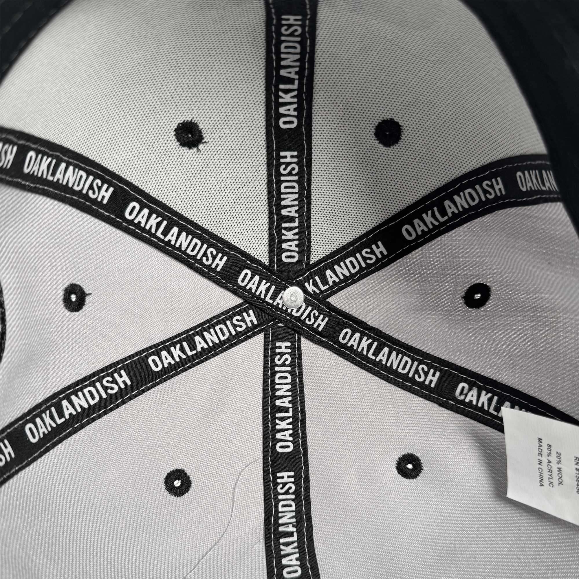 Close-up of the inside of the gray Oakland Roots SC Arch Text Snapback by Oakland Roots SC, featuring black fabric strips with repeated white "OAKLANDISH" text, black stitching and eyelets, and a partially visible white tag.