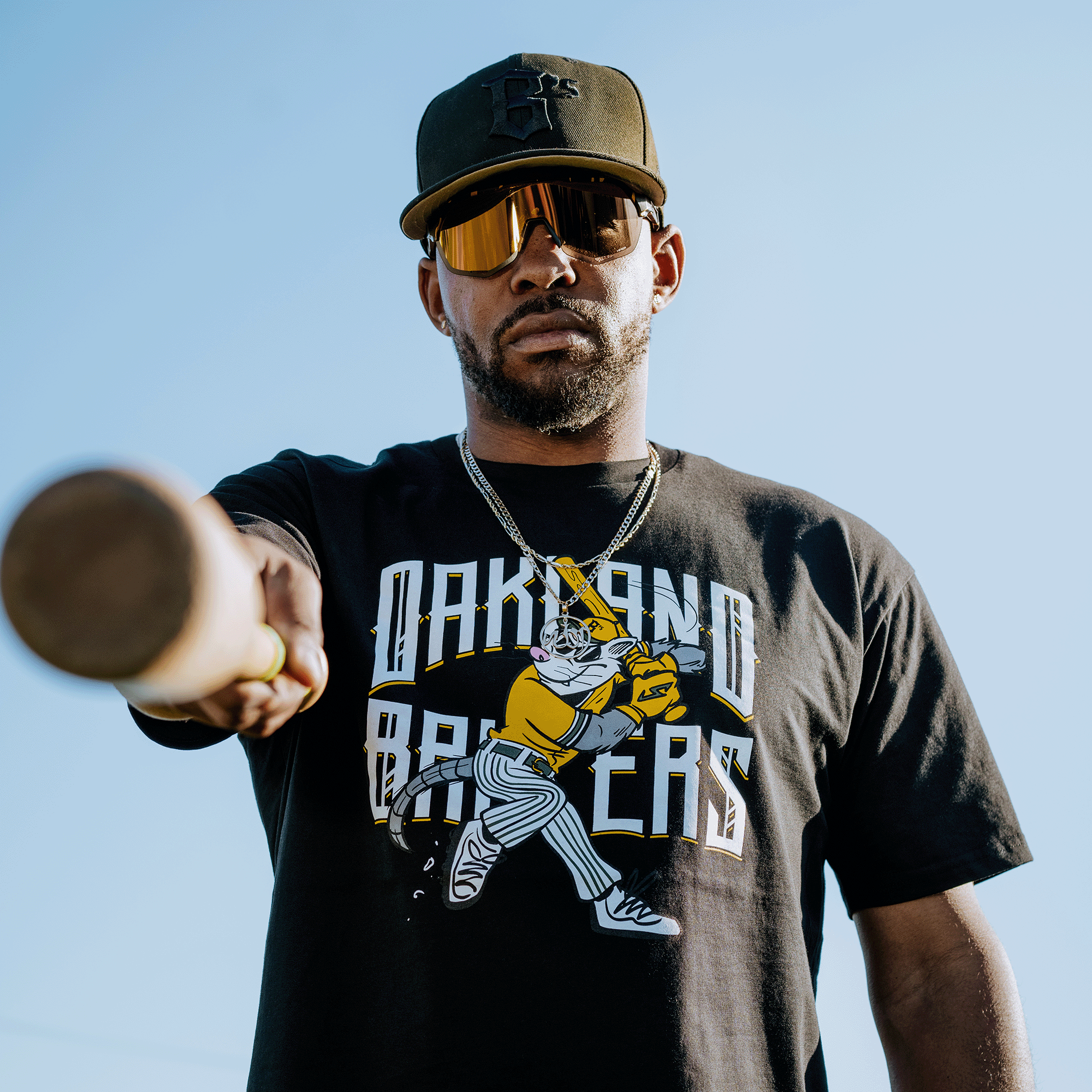 A man in a black cap and sunglasses poses with a bat, wearing the Oakland Ballers' Slugging Scrappy Tee featuring their mascot, Scrappy the Rally Possum. Against a clear blue sky, he completes his look with a gold chain necklace.