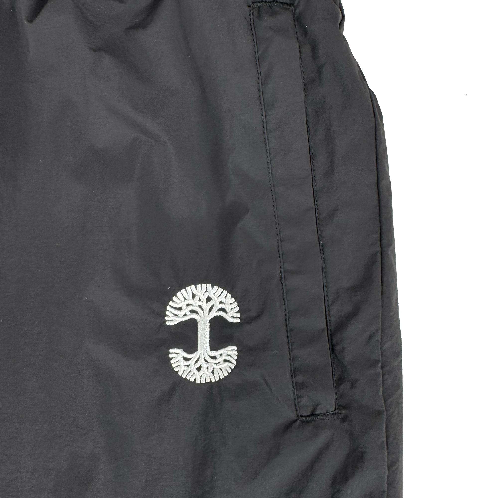 Close-up of the Oaklandish Susurrus Track Bottom in black nylon, featuring a white embroidered tree logo near the pocket, vertical zipper pocket opening, and visible stitching details. Set against a plain white background to highlight its modern style.