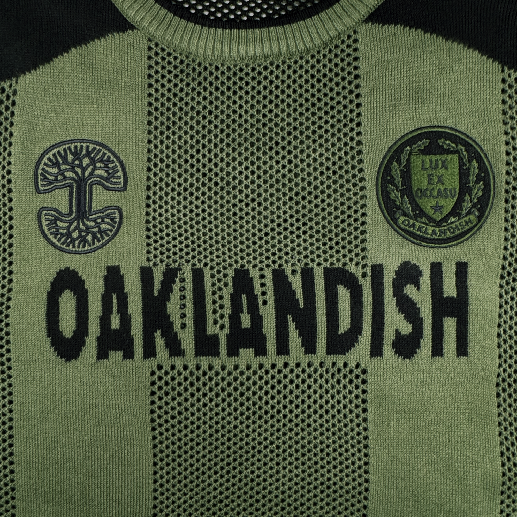 The Oaklandish Terrace Sweater by Oaklandish features a green and black knit pattern, bold "OAKLANDISH" lettering across the chest, and two army-style patches—a tree logo and luxe crest—designed in men's sizing.