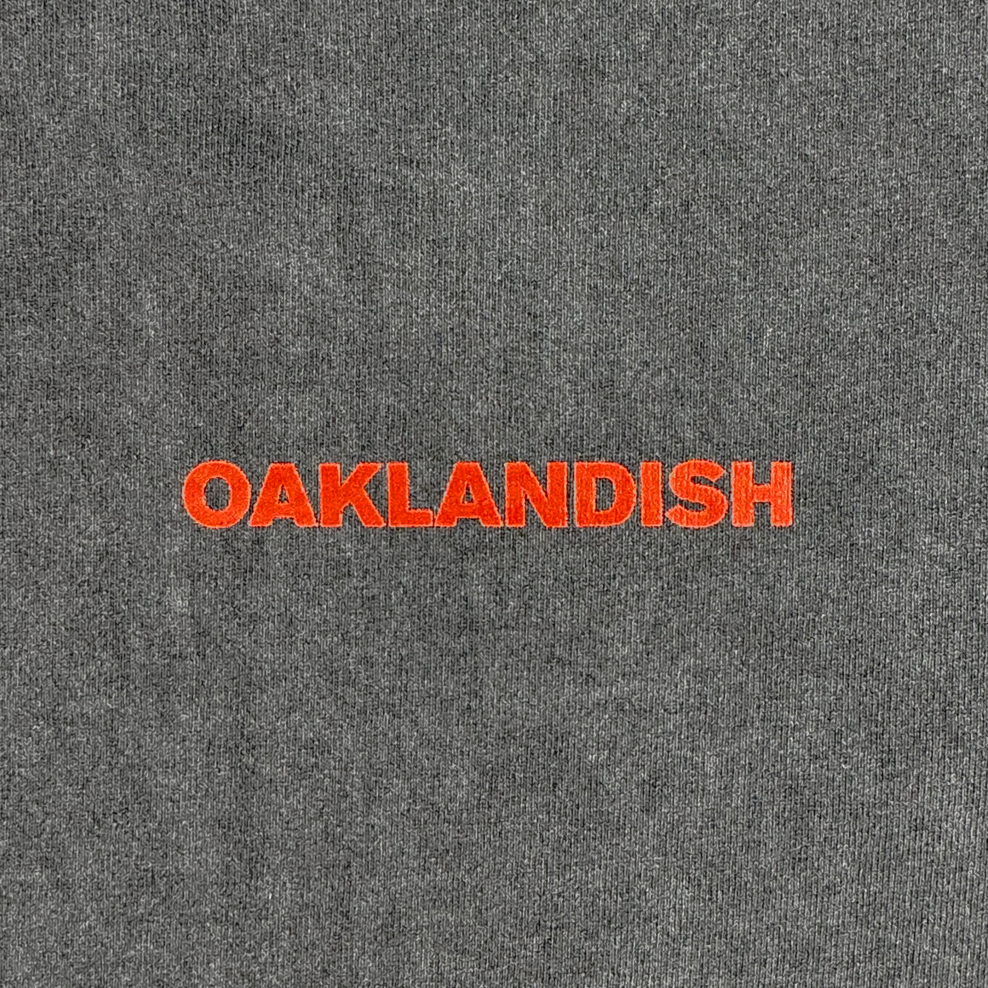 A close-up of the Tiger Blood Tee by Oaklandish shows gray, 100% cotton fabric with "OAKLANDISH" boldly embroidered in orange uppercase letters, centered on vintage black tee-like material against a plain background.