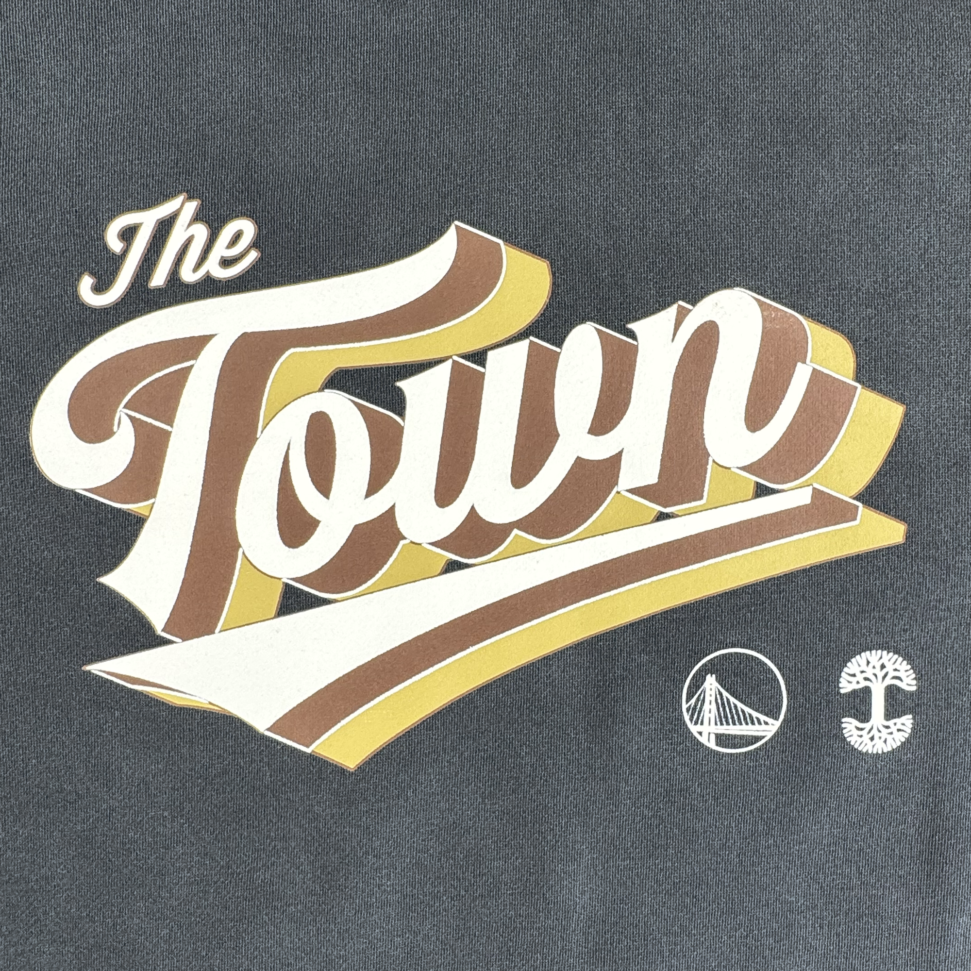 The GSW Vintage Swash Hoodie features stylized "The Town" text in cream, yellow, and brown on dark fabric. Below are white bridge and tree icons, a nod to Oaklandish roots. This retro design is part of the Warriors-inspired Town Collection.