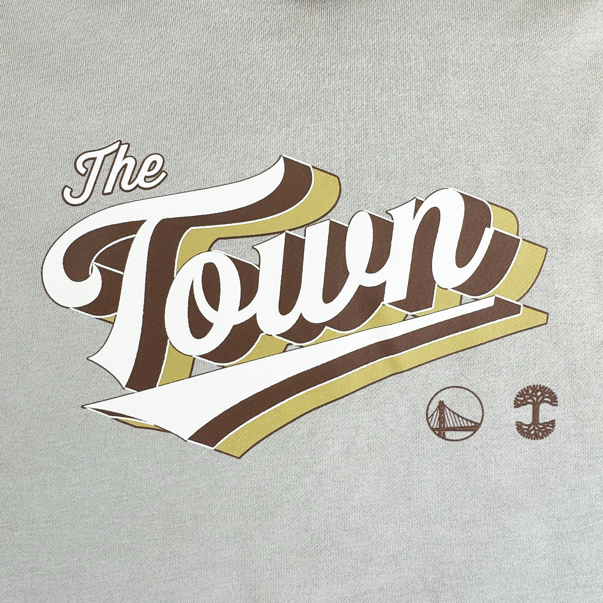 The GSW Vintage Swash Hoodie displays "The Town" in white, brown, and gold lettering with a bridge and tree icon below, all set on light gray fabric.