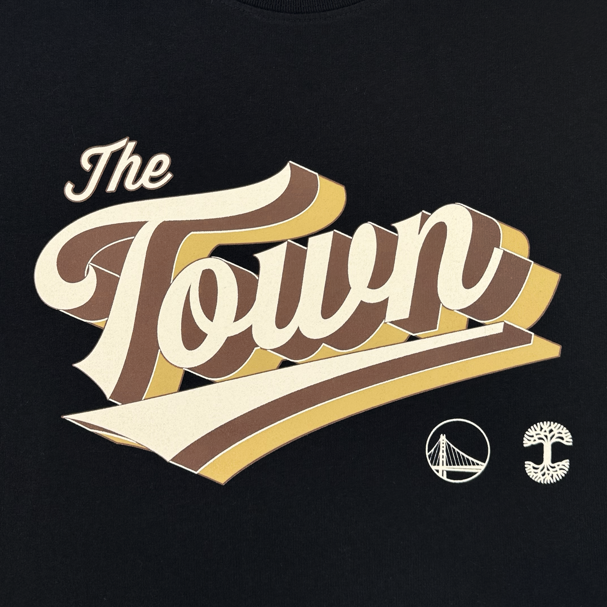 The Vintage Swash Tee by GSW features “The Town” in bold, retro-style script with brown, cream, and yellow stripes above the Warriors’ Bay Bridge logo and a stylized oak tree, inspired by Oaklandish’s Town Collection on black.