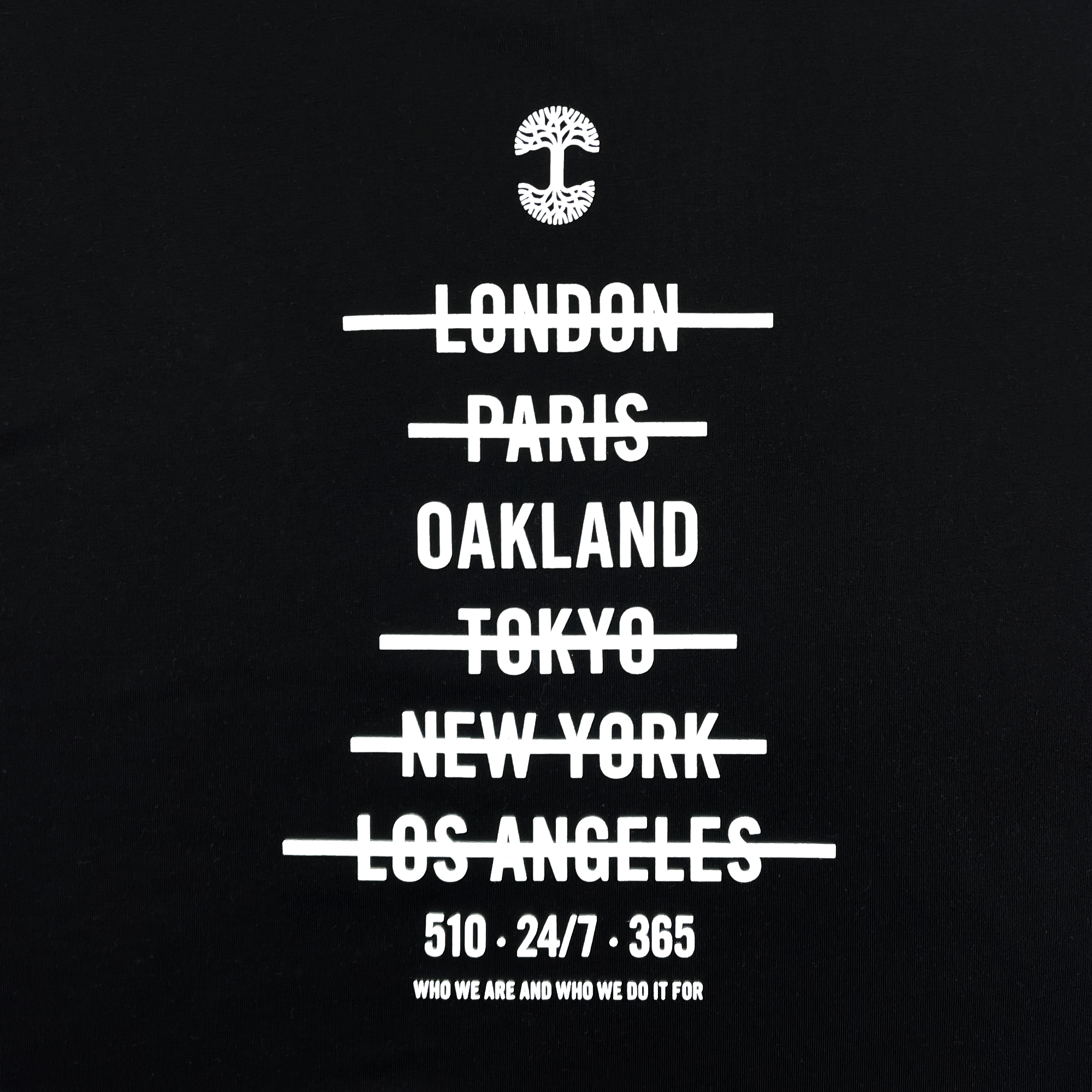 The "Who We Are and Who We Do it For Tee" by Oaklandish features a black background with white text. At the top is a tree logo. Below the logo, the names of cities—"London," "Paris," "Oakland," "Tokyo," "New York," and "Los Angeles"—are written and crossed out. Underneath these, on a classic fit t-shirt made of 100% cotton, are the phrases: "510 - 24/7 - 365" and "Who we are and who we do it for.