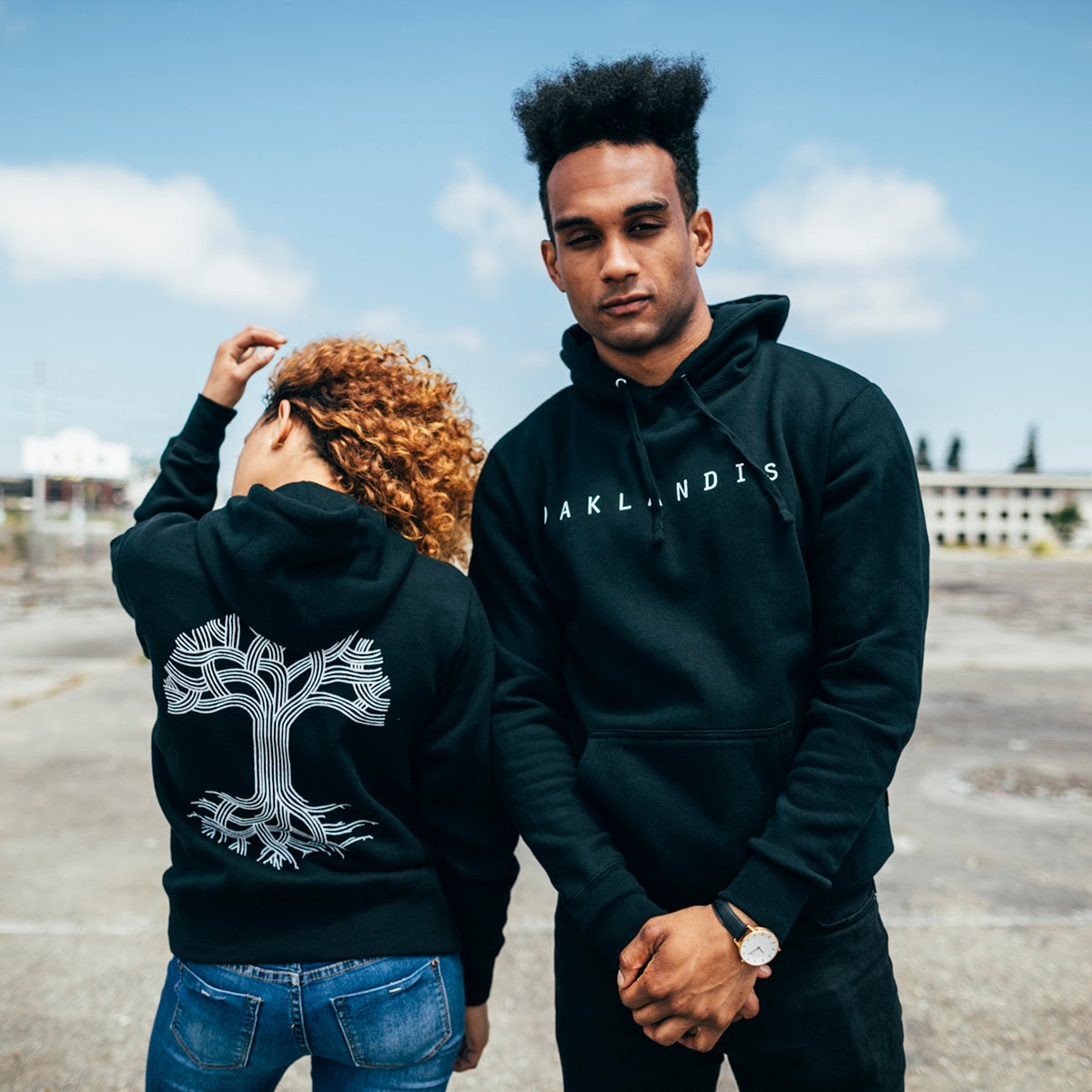 Two individuals are outdoors, each donning black Oaklandish Classic Hoodies. The person on the left turns away, revealing a stylized tree graphic on the back of their hoodie. The individual on the right faces forward with "Oaklandish" displayed on their men's fit hoodie. A clear sky and urban elements in the background highlight Oakland roots.