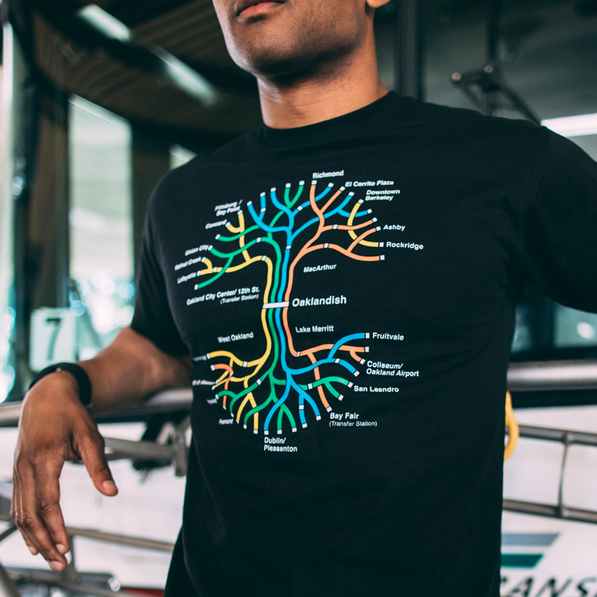 A person wearing an Oaklandish BART Tree Tee, a classic fit black T-shirt featuring a colorful transit map design, is seen in a casual setting. The vibrant map, resembling the BART system with various station names and lines, stands out on the shirt. The person is holding onto something off-camera with part of a bus or tram visible in the background.