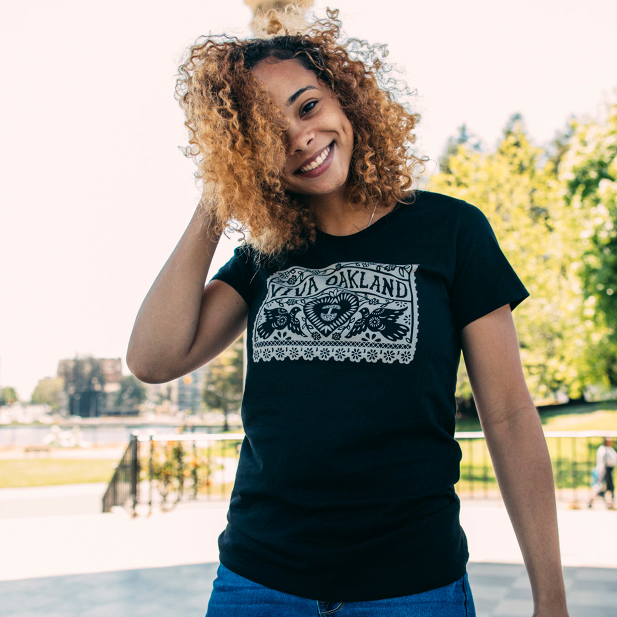 A woman with curly hair smiles at the camera while standing outside in a park. She is wearing an Oaklandish Women's Viva Oakland Tee, which is a 100% cotton, classic fit T-shirt featuring the word “Oakland” printed in white. Trees and a building are visible in the background.
