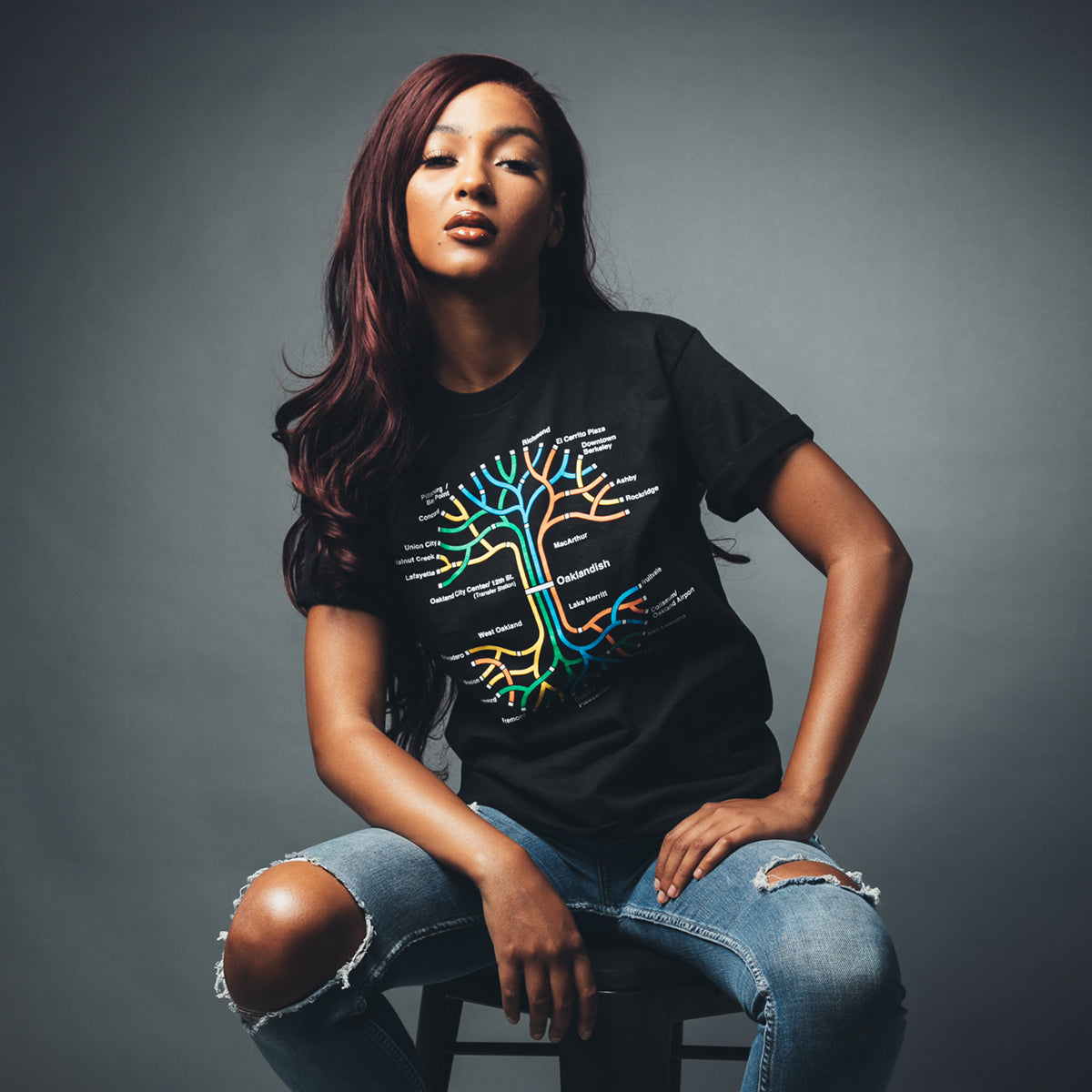 A woman with long, wavy auburn hair sits confidently on a stool against a gray background. She's wearing Oaklandish's BART Tree Tee, a classic fit black T-shirt featuring a colorful tree design with various words on its branches, paired with ripped blue jeans. Her expression is self-assured, with her head slightly tilted.